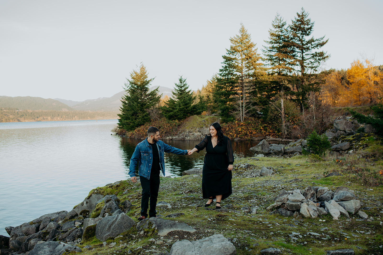 man holds hands with woman along shore with trees in background