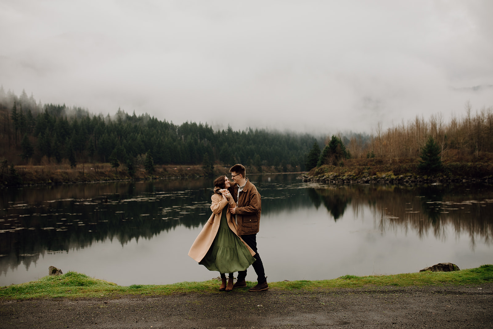 a man and woman stand at the bank of a lake with a cloudy forested mountain in the background reflected in the water