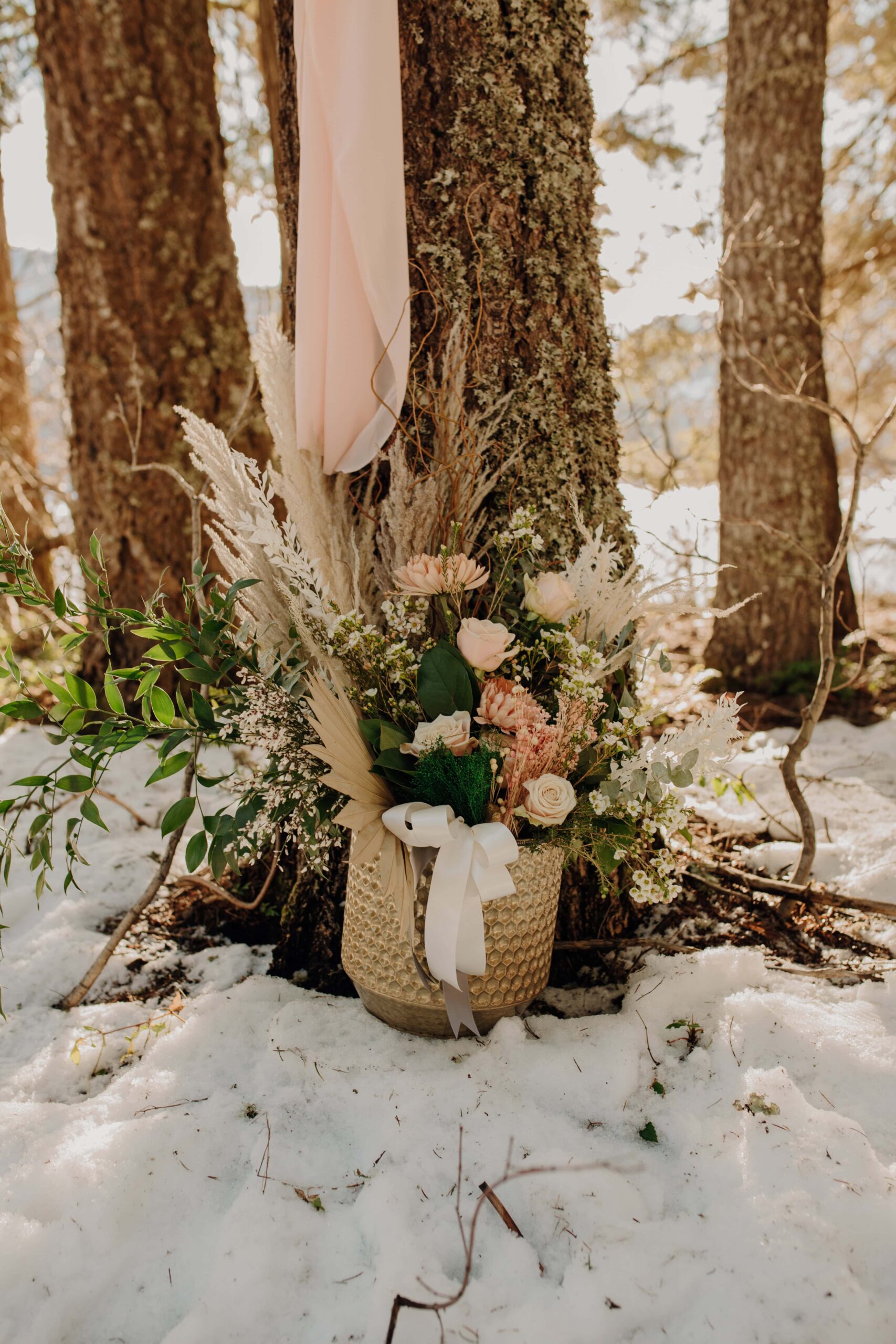 a basket of pink flowers and pampas grass leans against a tree in the snow for a Winter elopement at Lake Easton in Washington