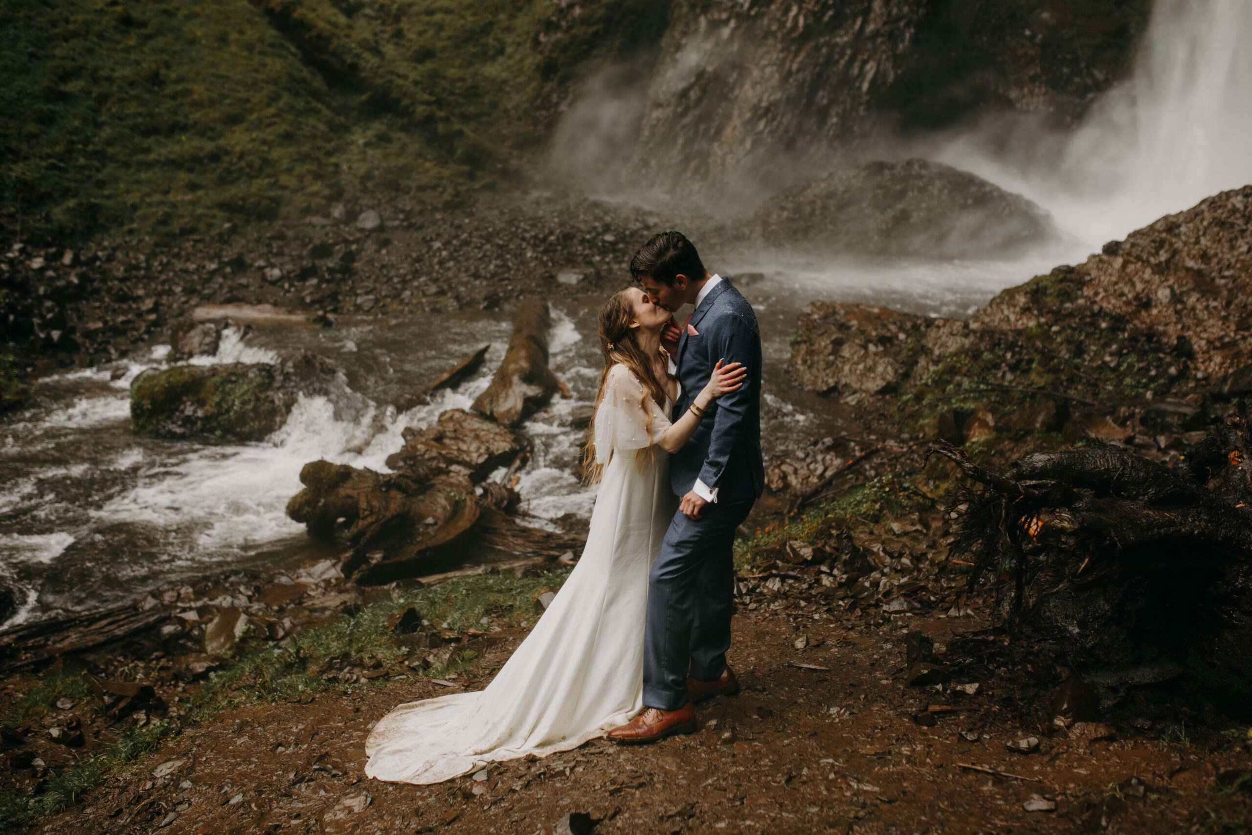 woman in white dress kisses man in blue suit outside near a waterfall on rocky shores