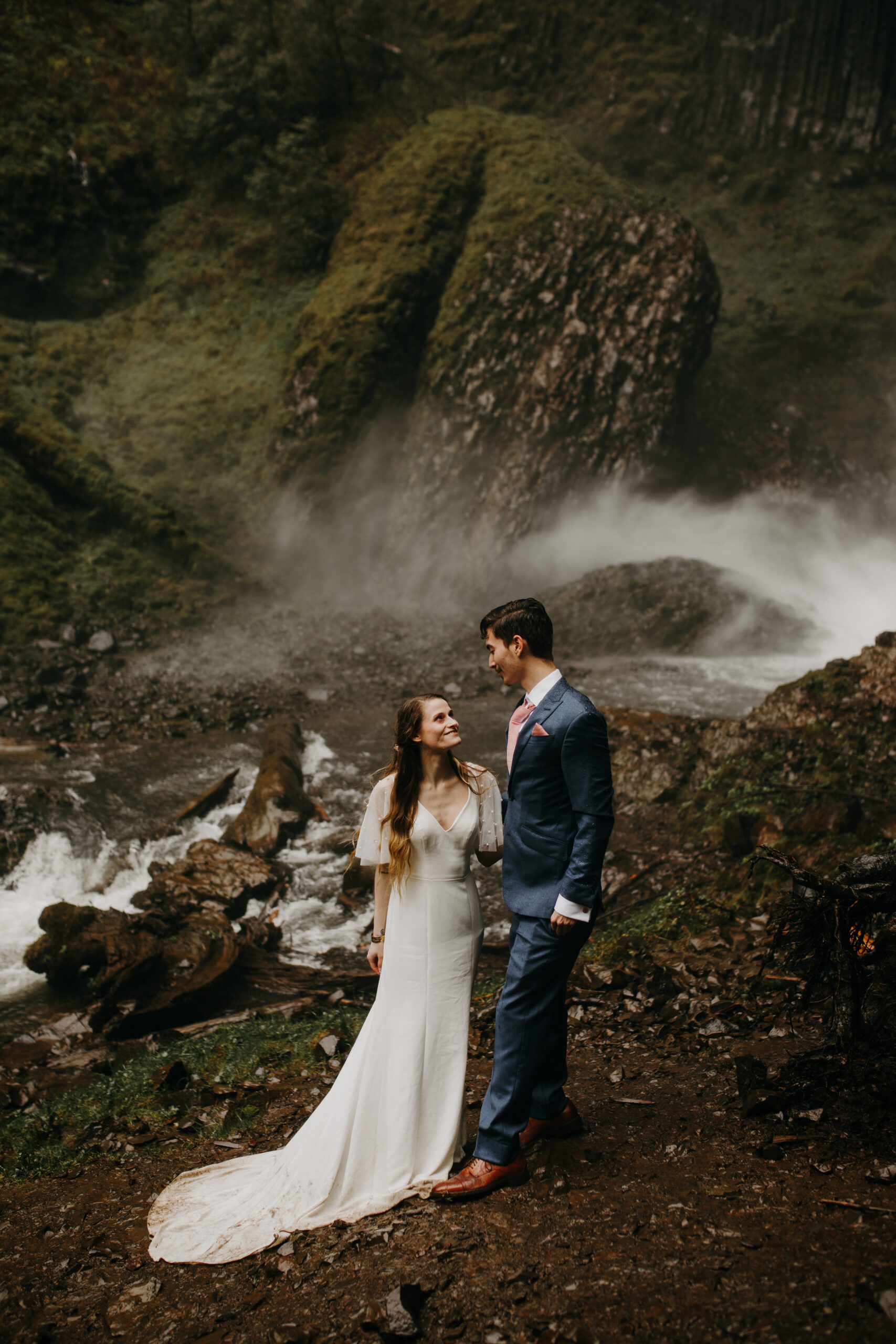 woman in white wedding dress stares smiling at man in blue suit and pink tie on a rocky shore near a waterfall basin. Latourell Falls elopement at Columbia River gorge. 