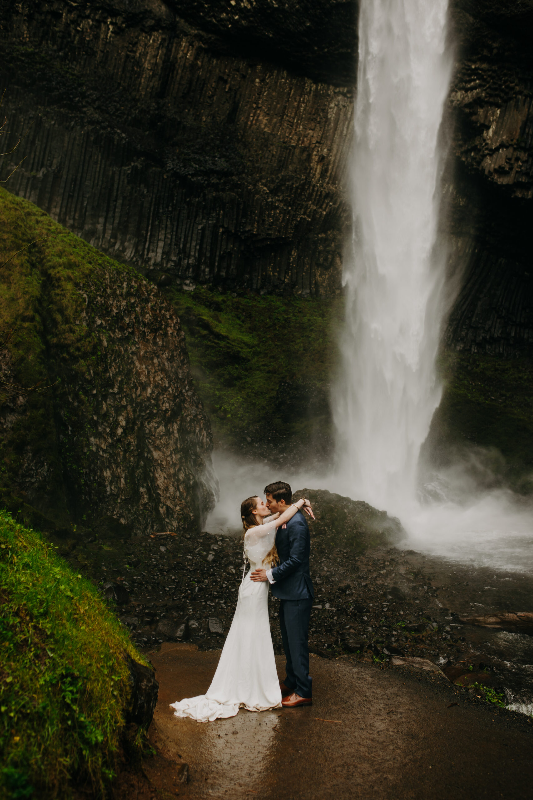 Woman and man embrace, woman is in wedding dress and man is in blue suit. They are standing outside in front of a waterfall. Latourell Falls elopement at Columbia River gorge.
