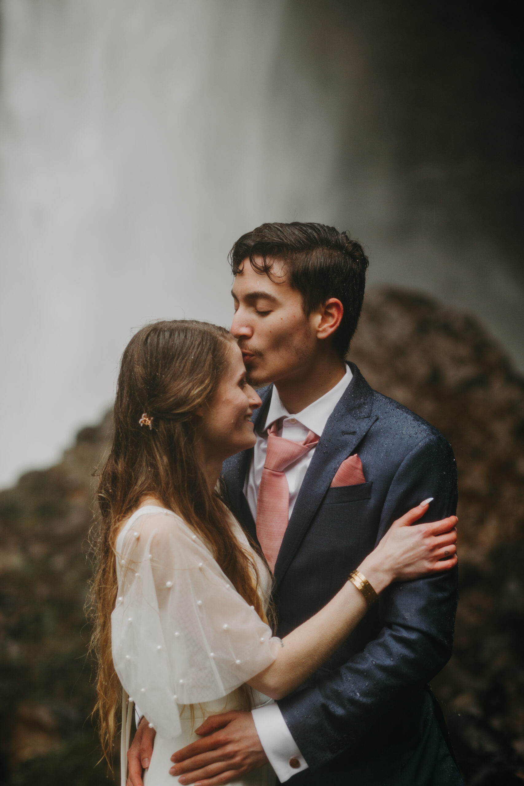Man in blue suit with pink tie kisses woman in wedding dress on the forehead with a waterfall in the background behind them. Couples session at Columbia River Gorge's Latourell falls