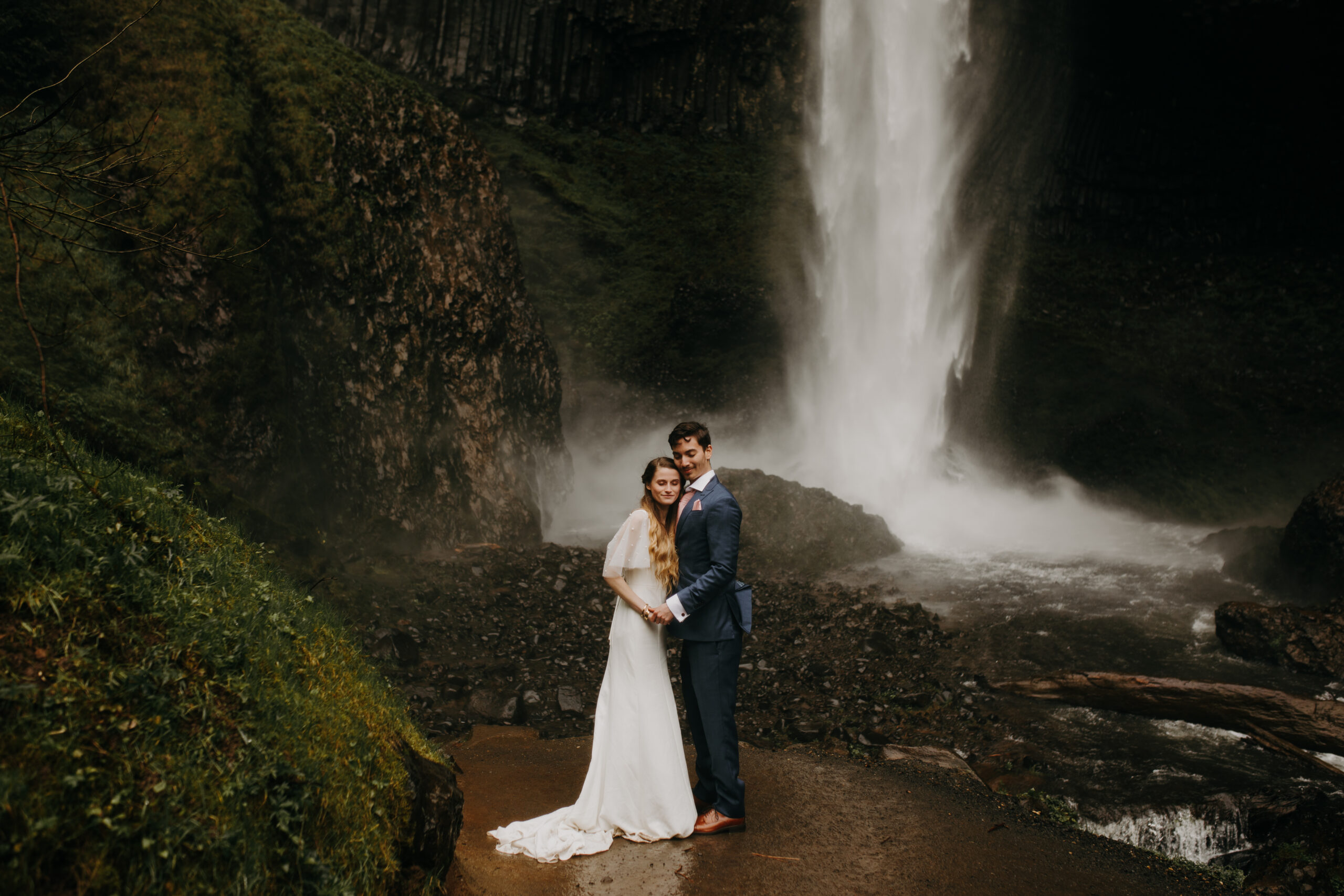 woman in wedding dress and man in blue suit with pink tie hold hands and smile with their eyes closed in front of Latourell falls in the columbia river gorge. 