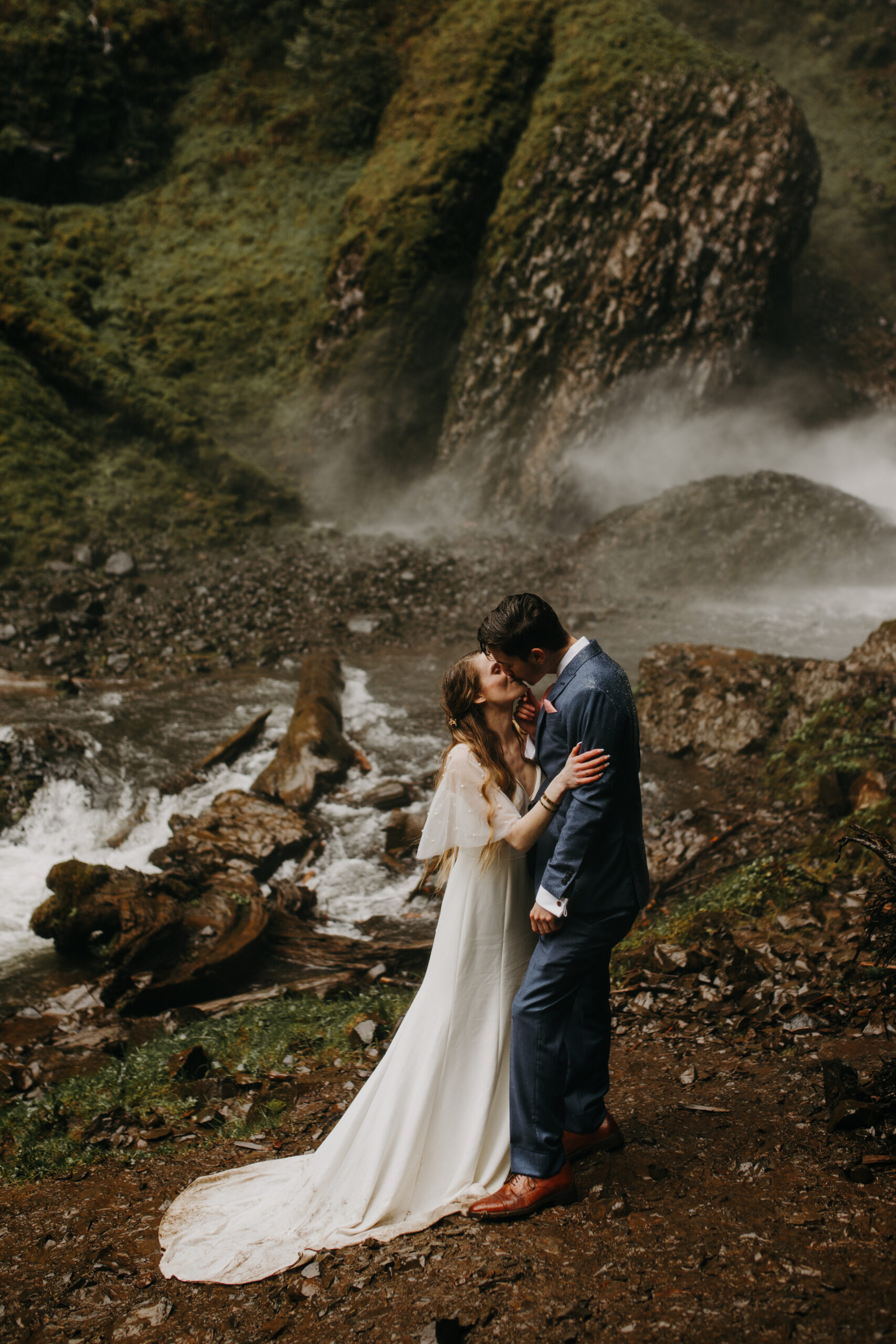 Woman in white dress kisses man in blue suit on rocky shores at Latourell falls in the Columbia River Gorge