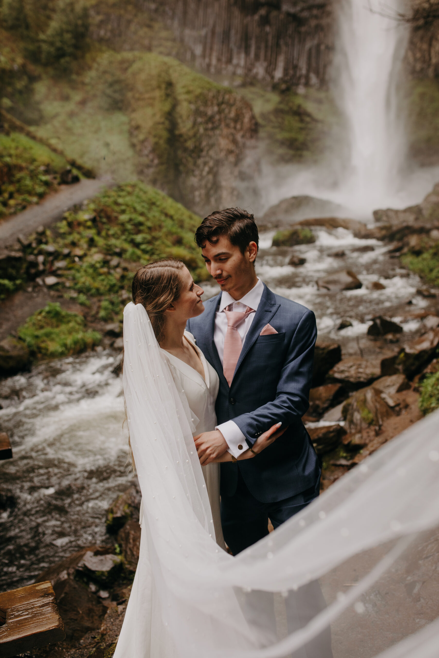 woman in white wedding dress and veil looks lovingly at man in blue suit at Latourell Falls in the Columbia River Gorge. 
