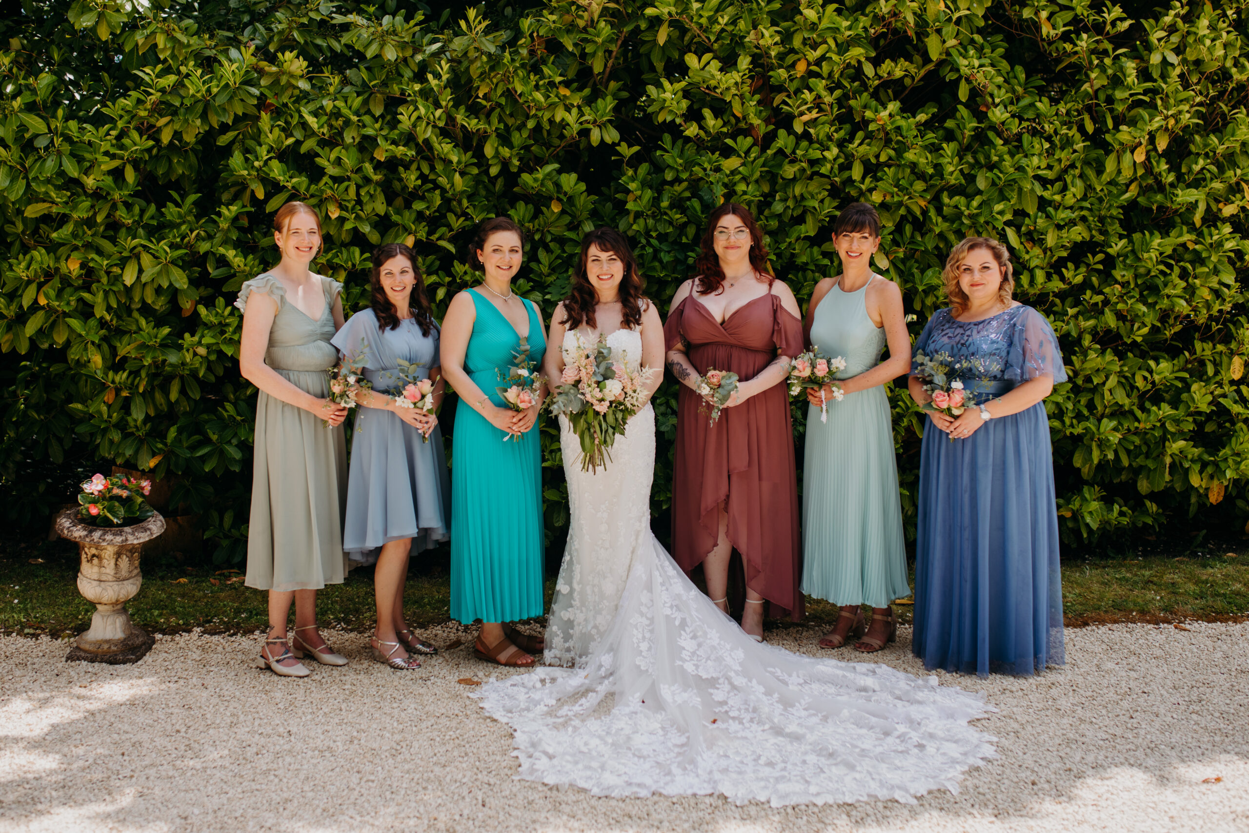 women in different dresses gather outside in front of ivy wall with bride in center
