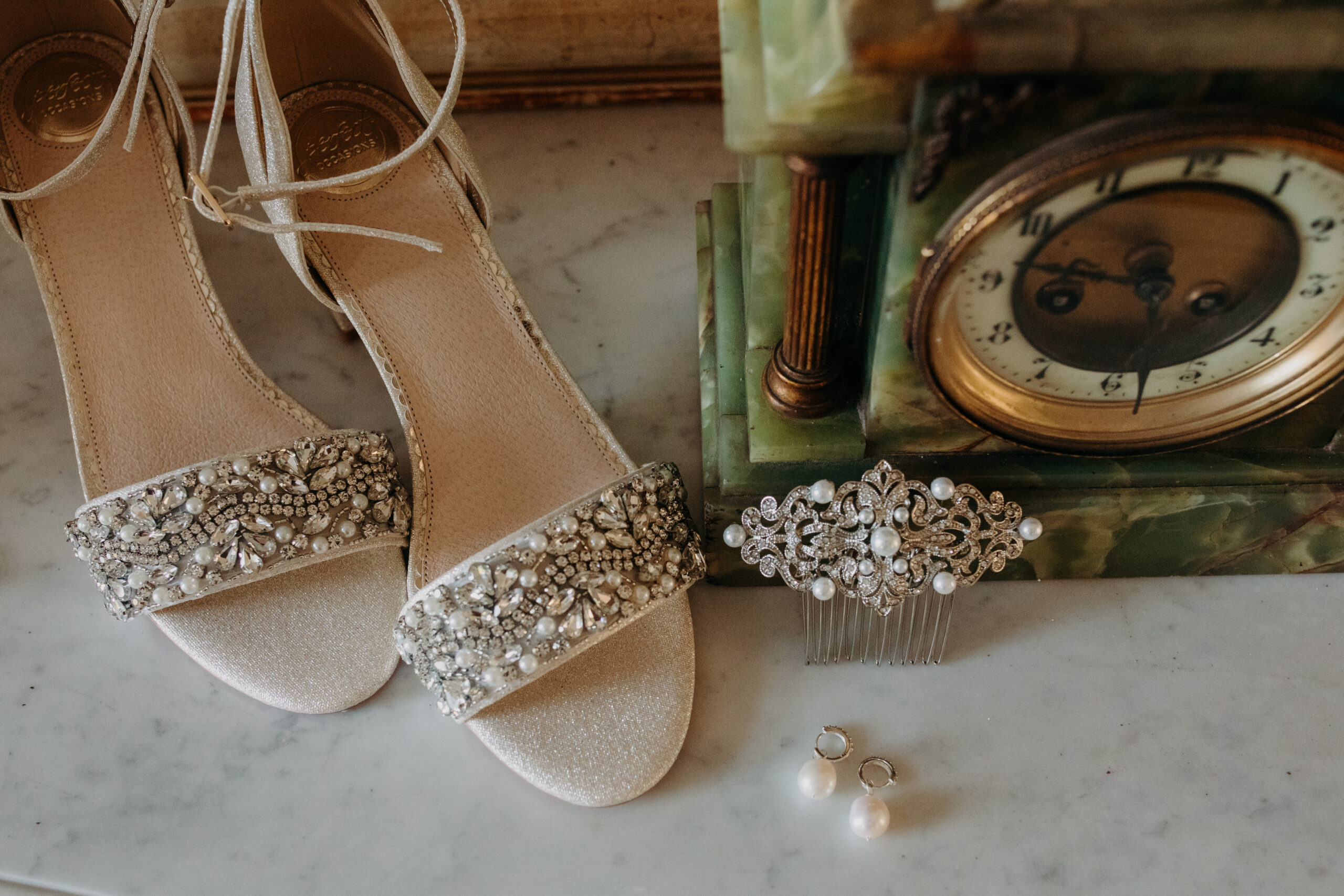 A bride's wedding heels sit to the left with a hair pin and earrings up against an old clock