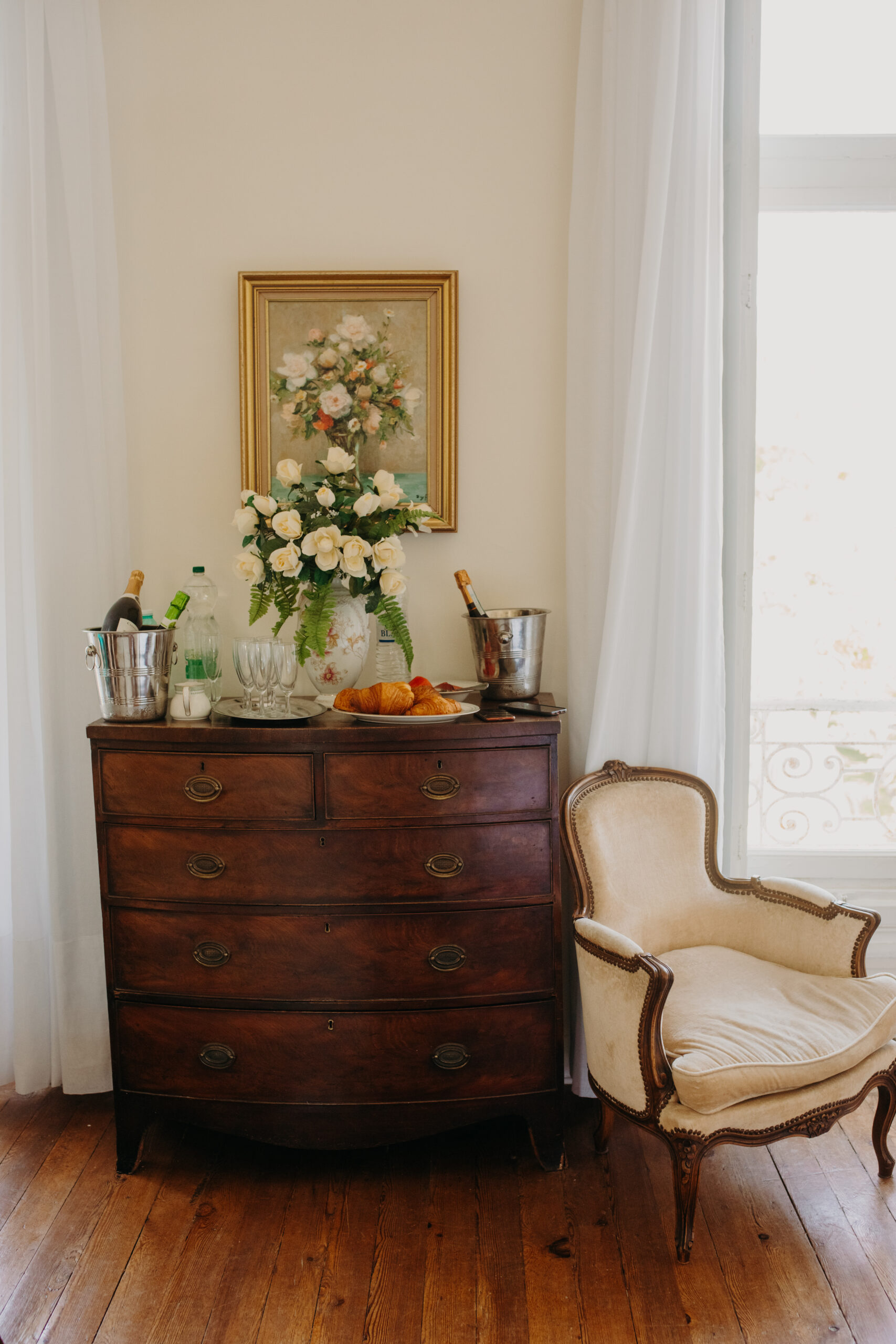 On a chest of drawers sits a flower bouquet with champagne in an ice bucket and croissants. 