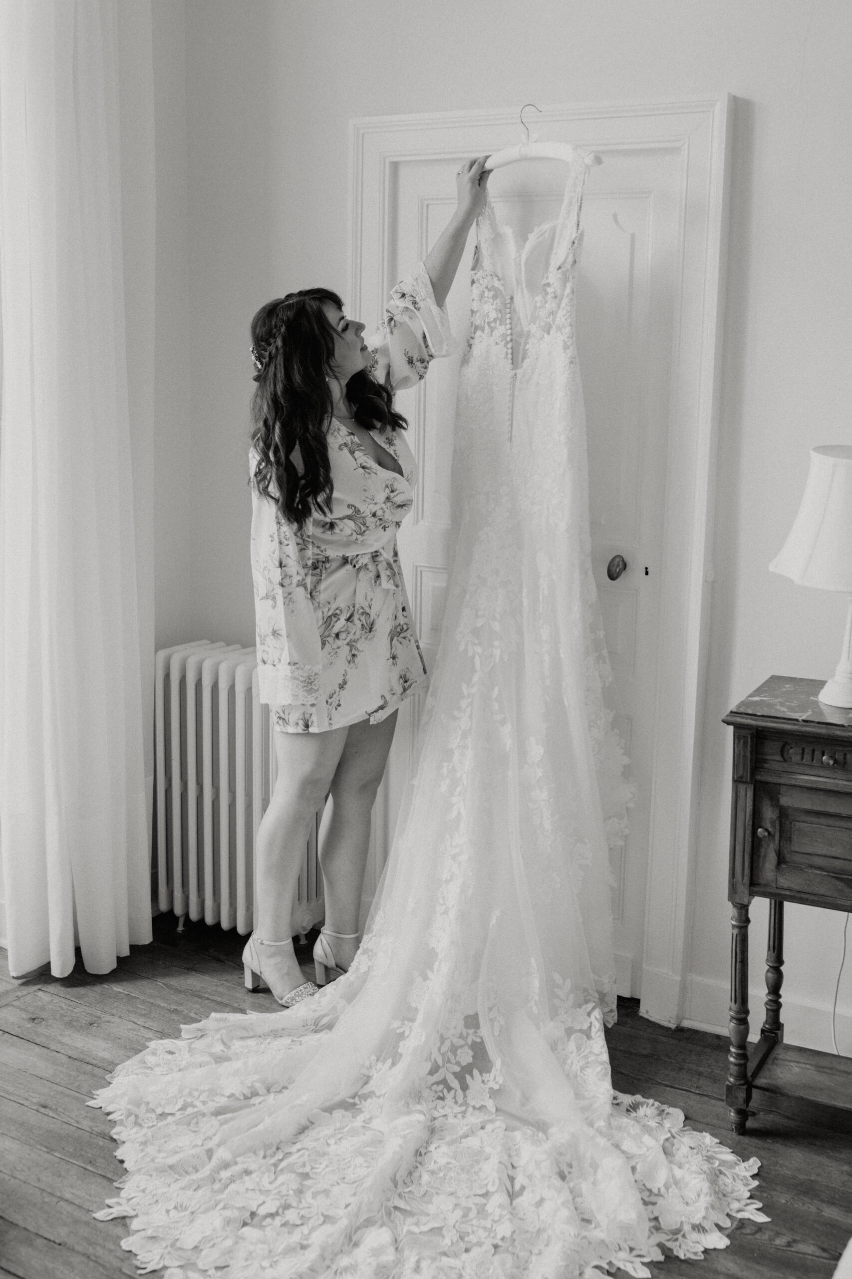 A woman in a robe reaches up to grab a lacy wedding dress hanging from a door.