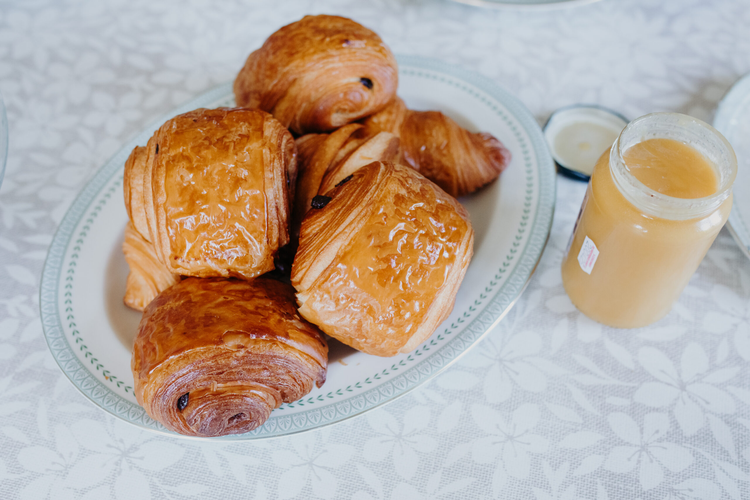Chocolate croissants sit on a white plate on a table with a white tablecloth and orange jam to the right. 