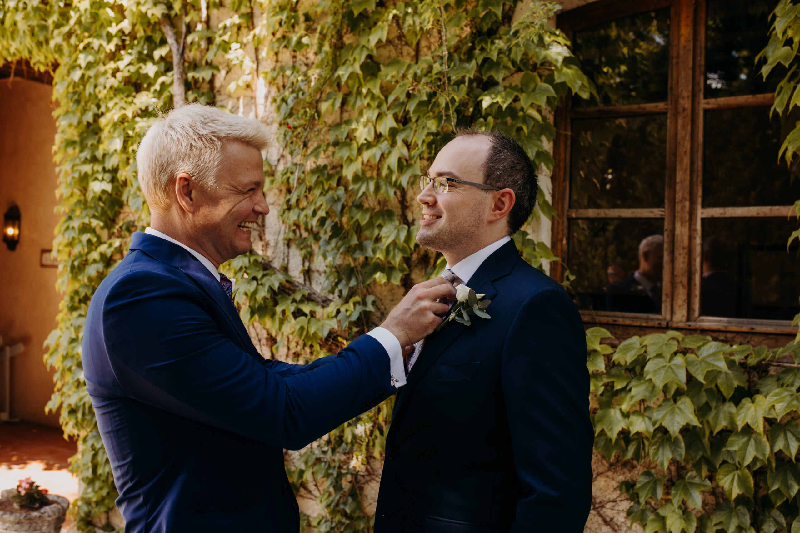 A best man with white hair adjusts the tie of a groom outside about to get married. both are smiling. 