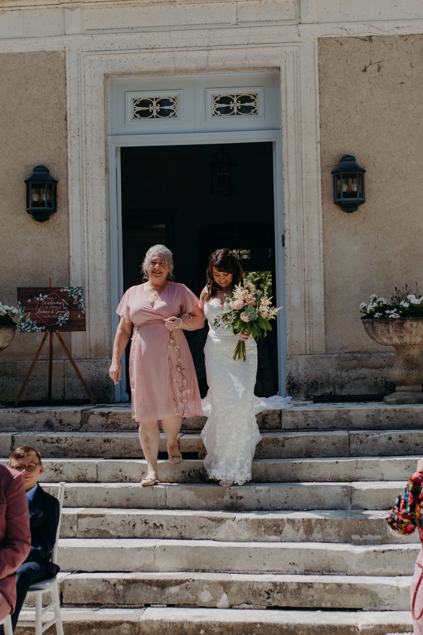 A woman in a pink dress walks a bride in a white dress happily down stairs