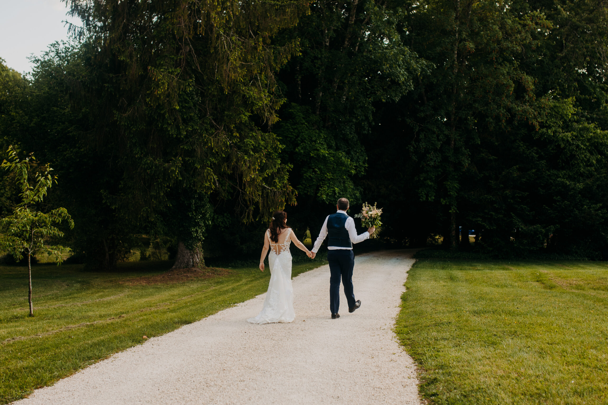 bride and groom face away from camera and walk down gravel path with the groom holding the bouquet