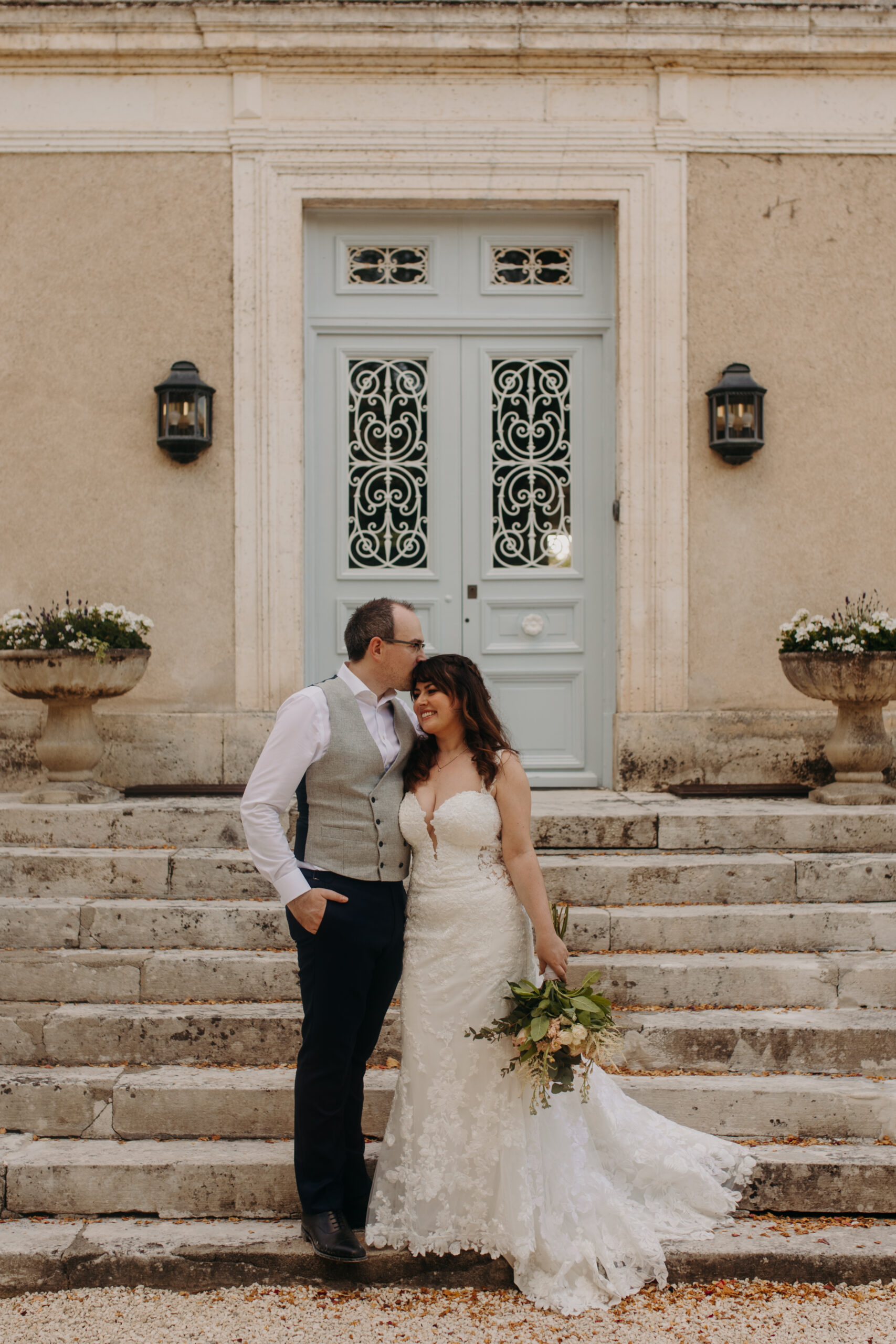 bride and groom stand on steps of chateau in front of blue door as groom kisses bride's head