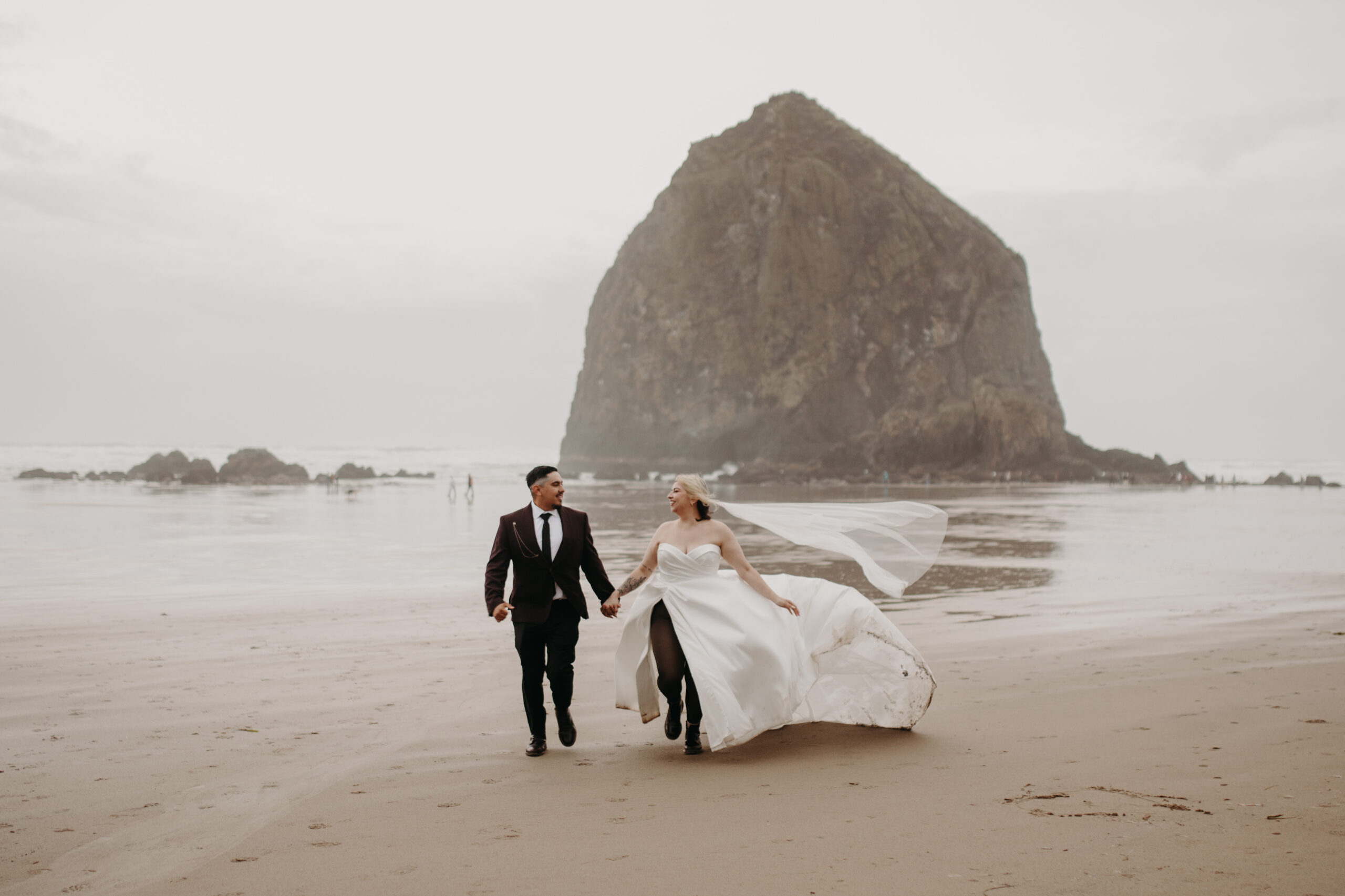 man in a suit and woman in a wedding dress run on beach smiling with haystack rock in the background