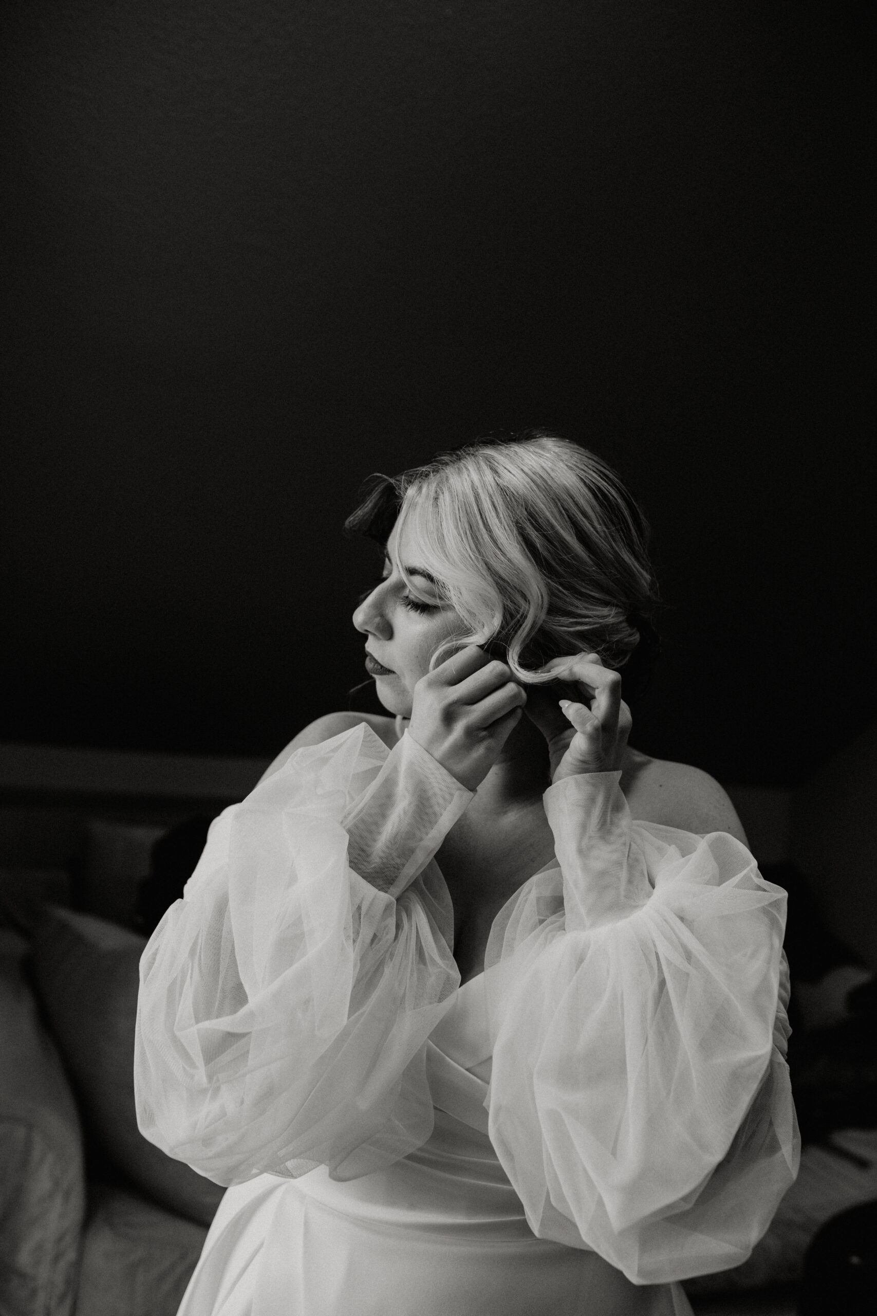 black and white photo of woman in white wedding gown putting earring on 