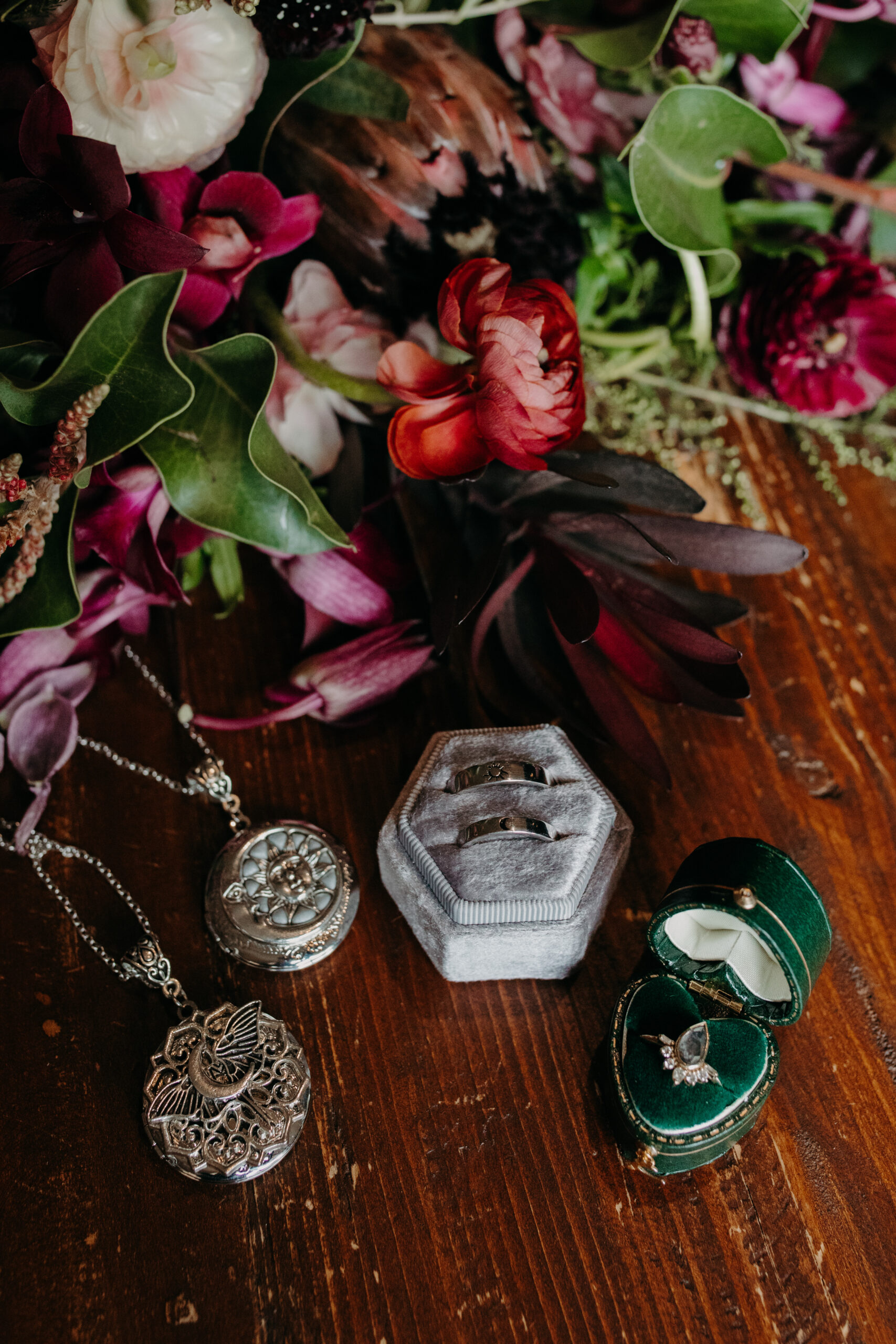 flat lay on wooden table of two silver lockets next to a grey ring box with two rings inside and beside a green heart shaped ring box with a ring inside 