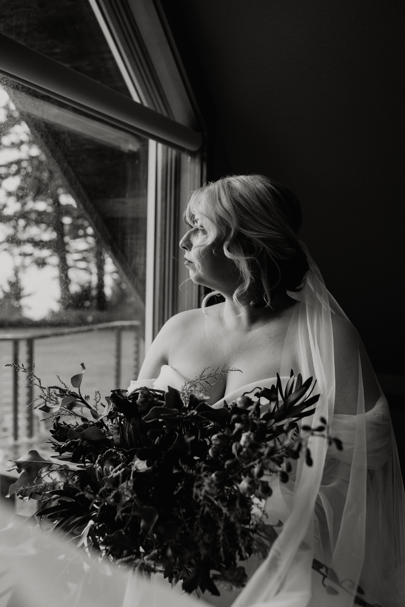 black and white photo of a bride holding bouquet of flowers looking out window 