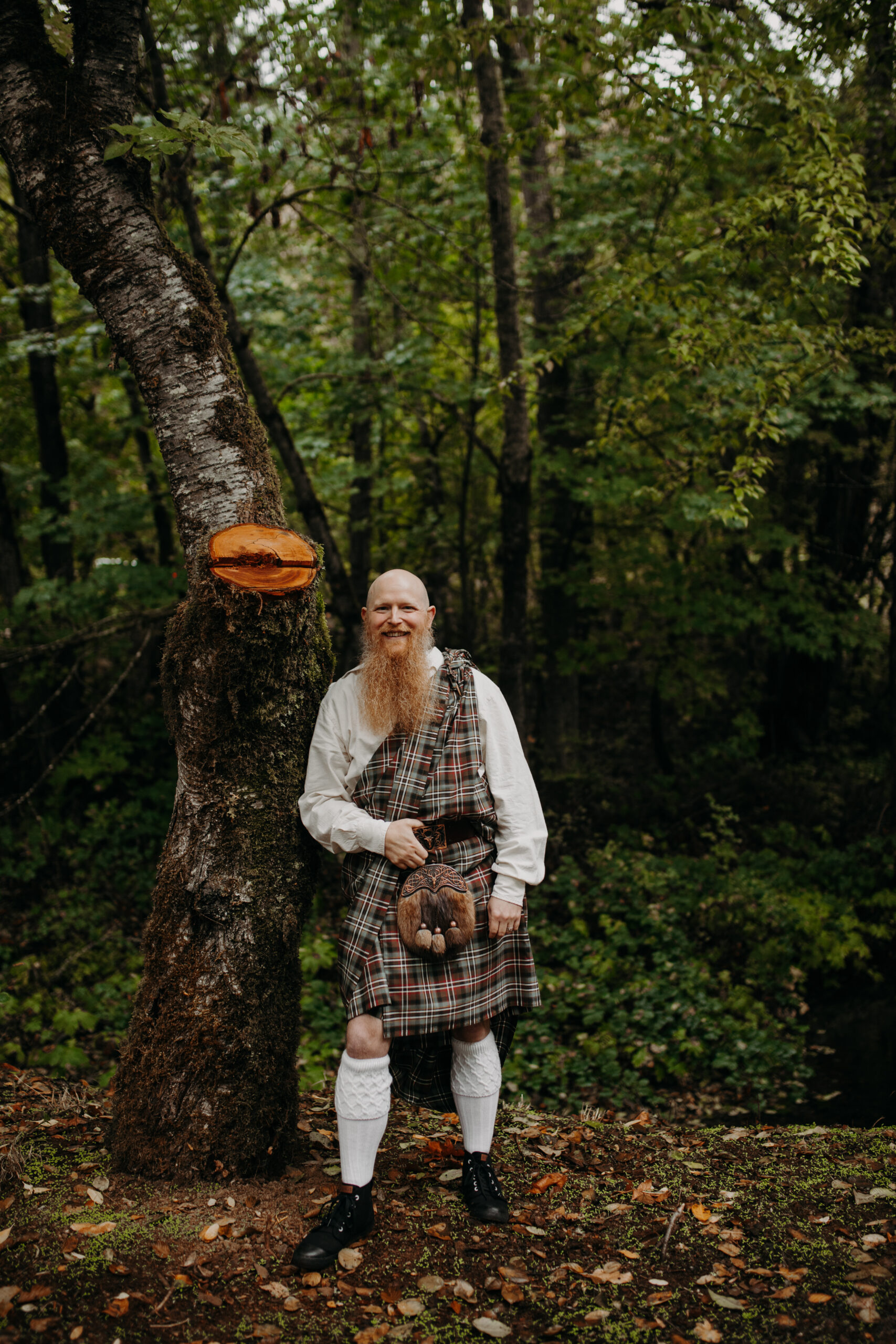 man in kilt leans against tree in a forest