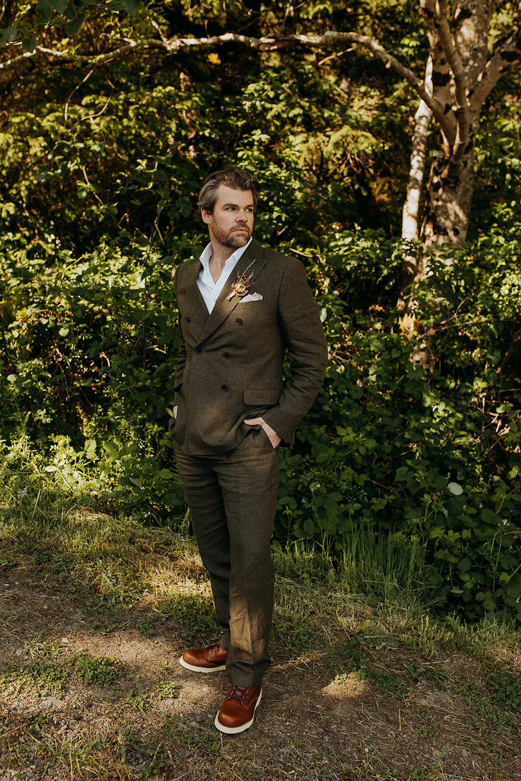 A groom stands in a dark green suit looking off into the distance with forest behind him for this Brookings, Oregon Elopement.