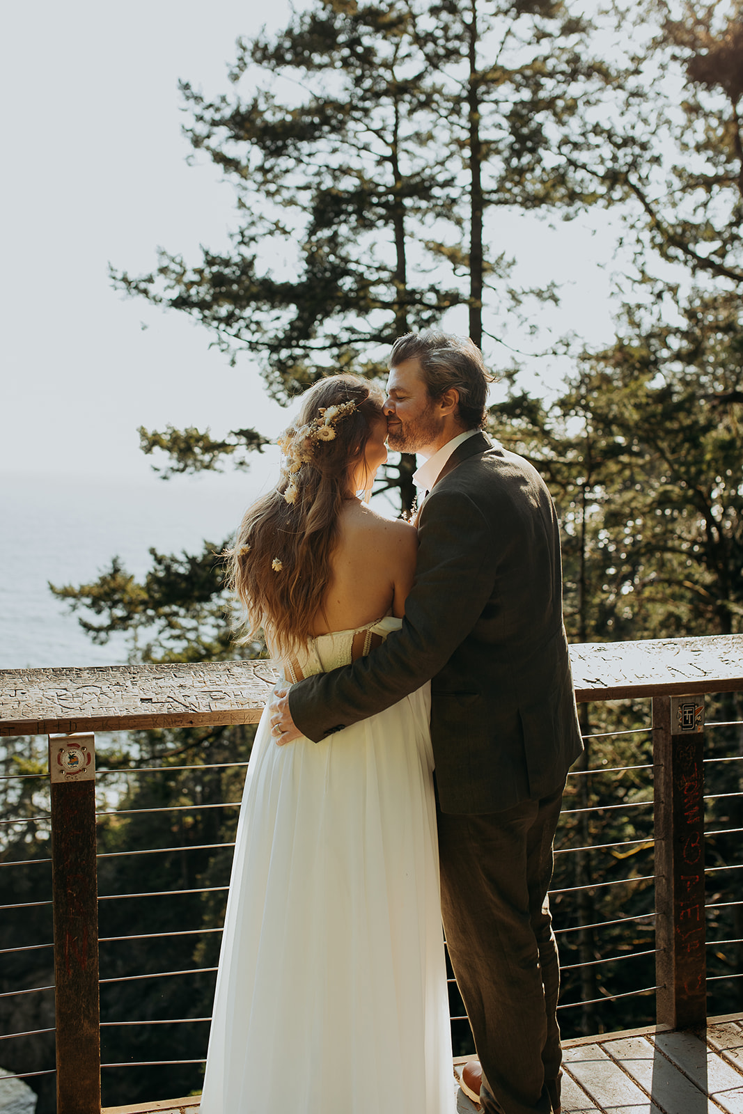 A couple stands before a railing as the bride looks out to the ocean with the groom smiling at her forehead