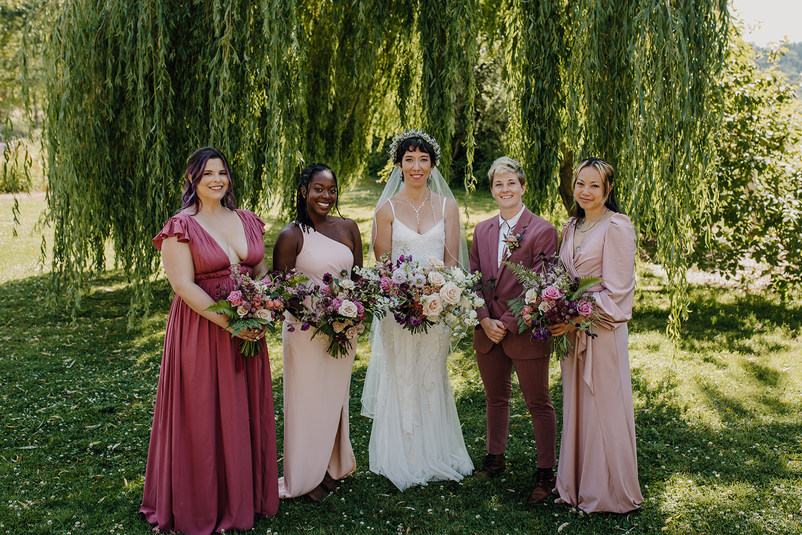 a group of bridesmaids stand in front of a weeping willow in semi-formal dress in shades of pink and mauve