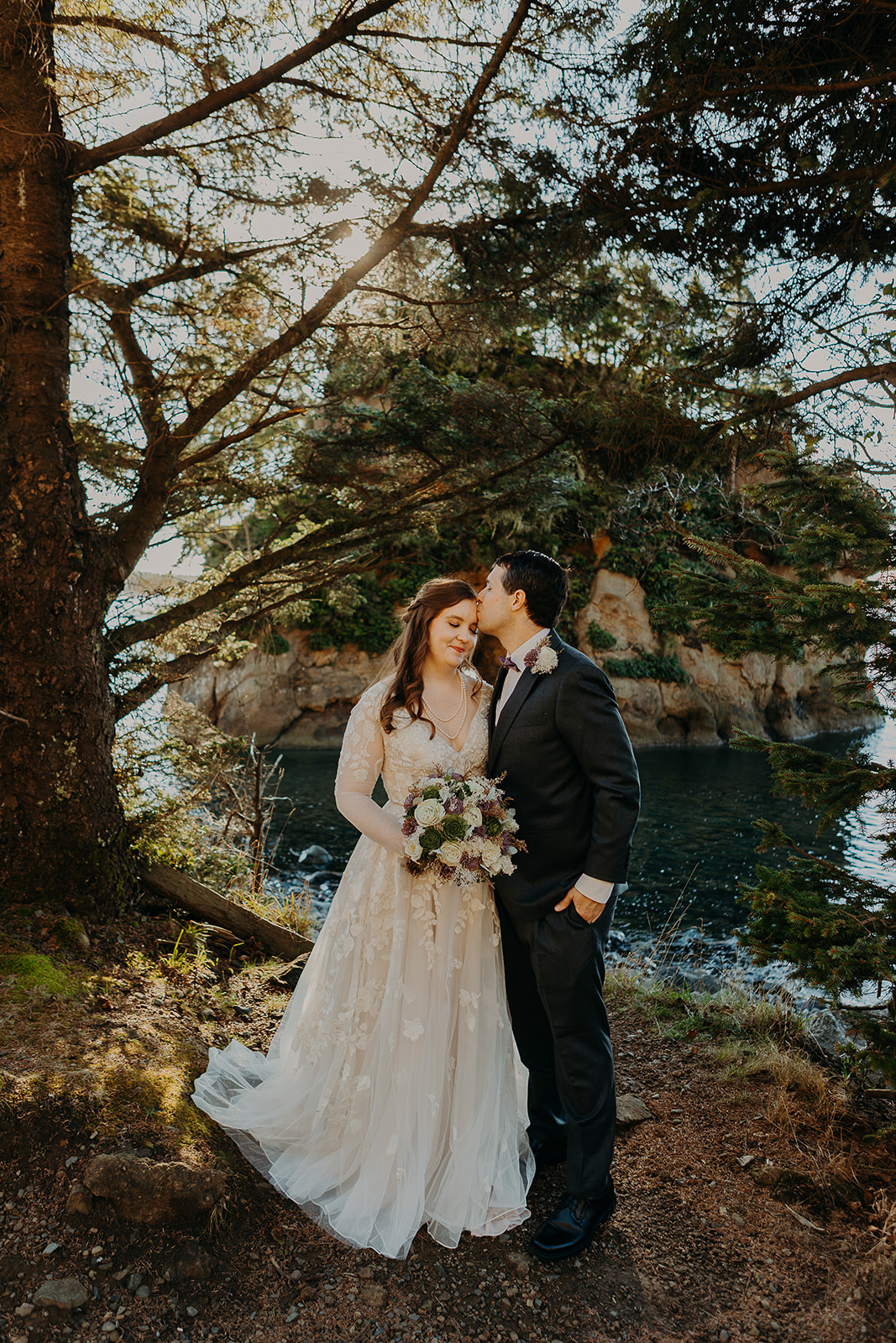 a couple in wedding attire stand in Rockaway Beach in front of a scenic rock with the ocean in the background