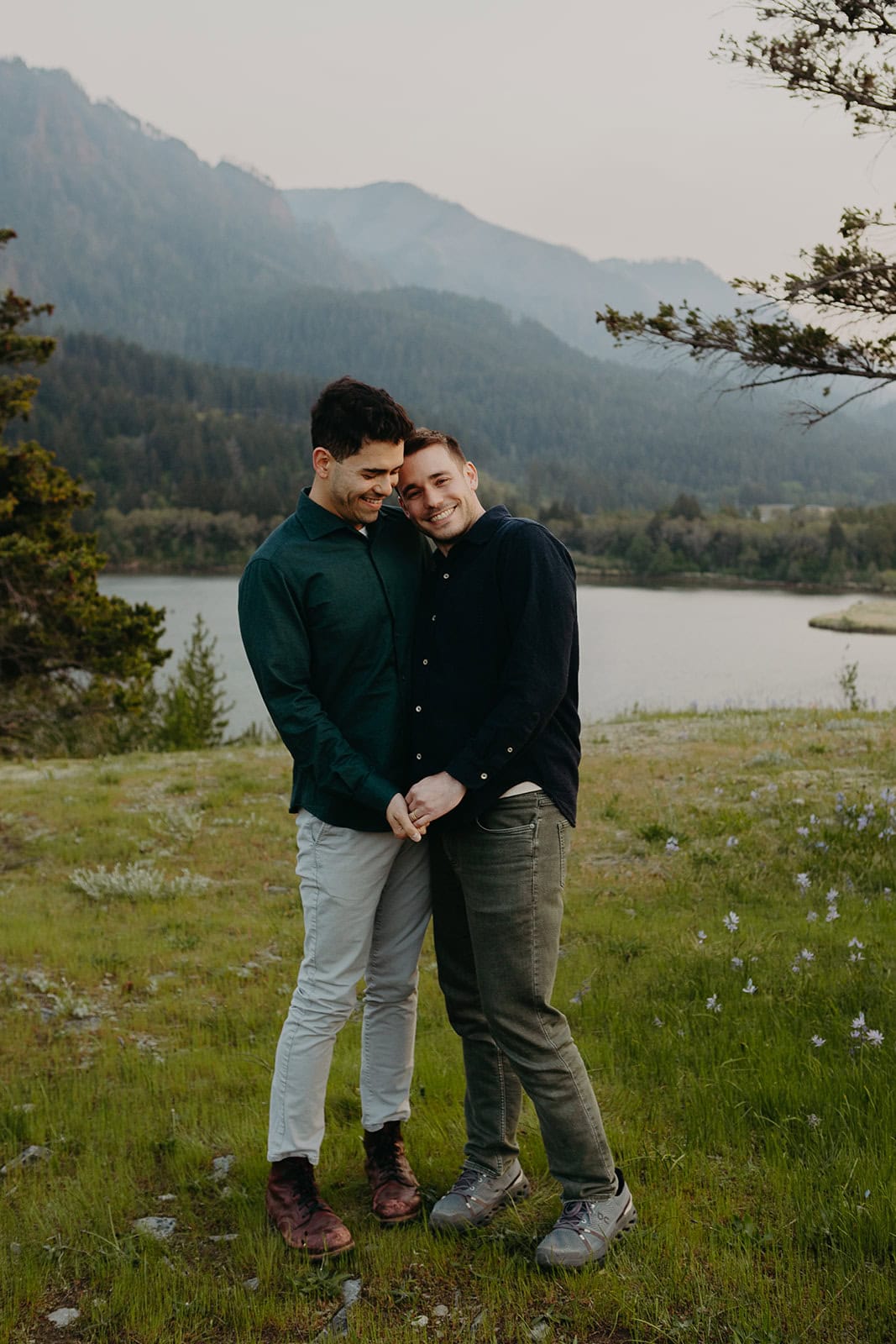 Two men in casual dress attire stand hand in hand as one leans into the other smiling amidst nature