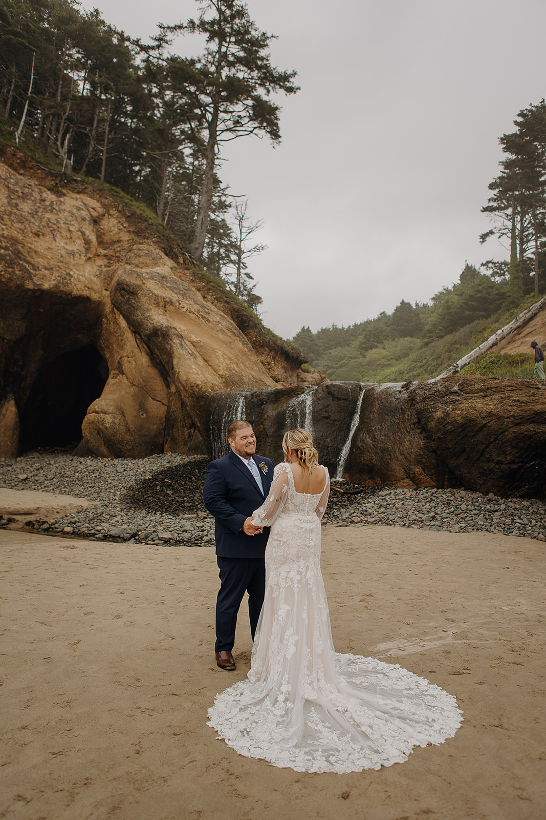 a man and woman in wedding gear stand smiling in front of the waterfall at Hug Point beach in Oregon. 