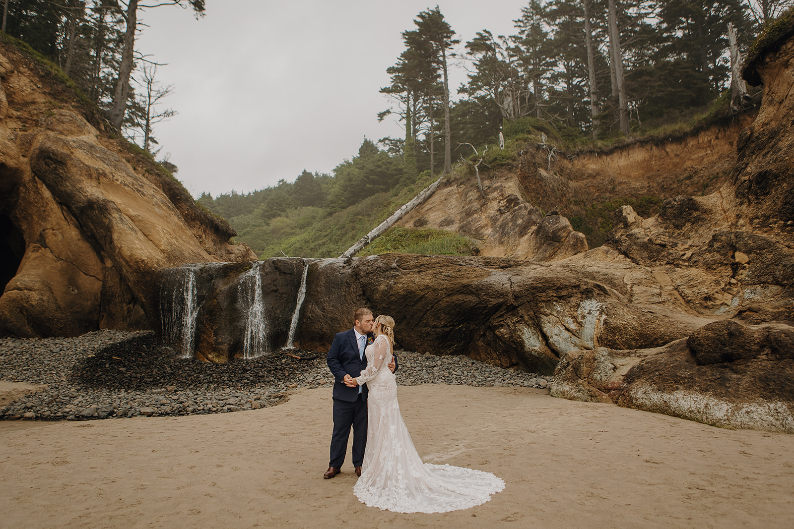 A couple in wedding outfits kiss in front of the waterfall at Hug Point State Recreation site. 