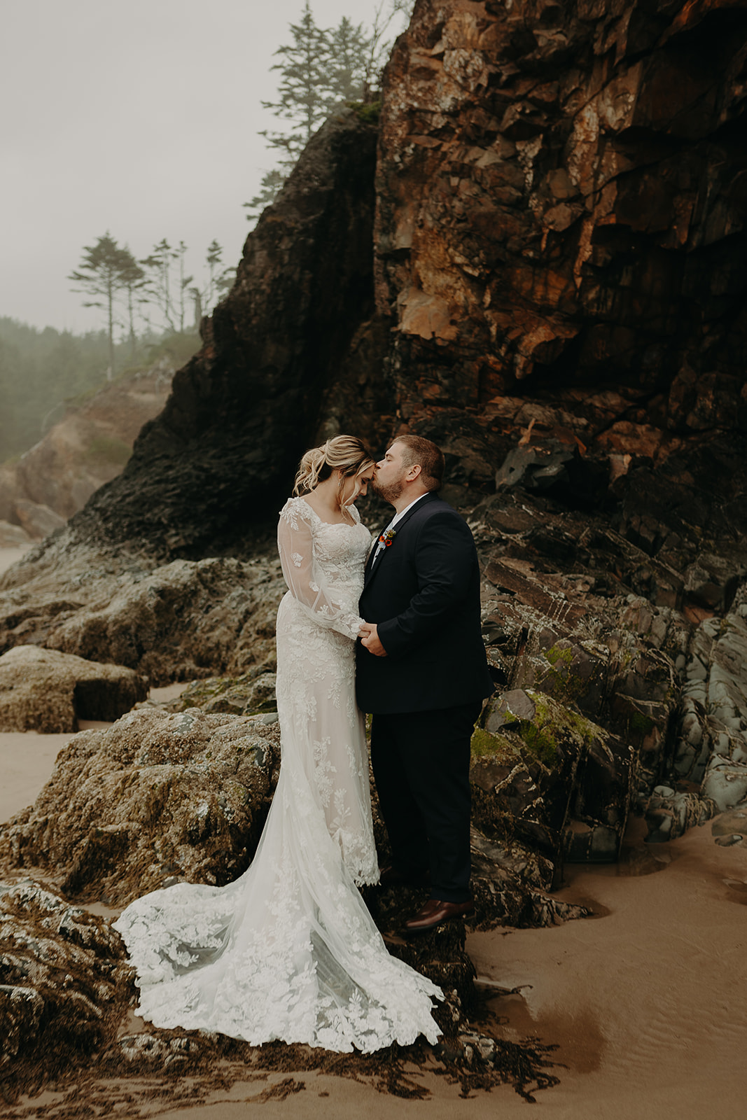 a couple in wedding gear stand on rocky shores at Hug Point State Recreation Site. 