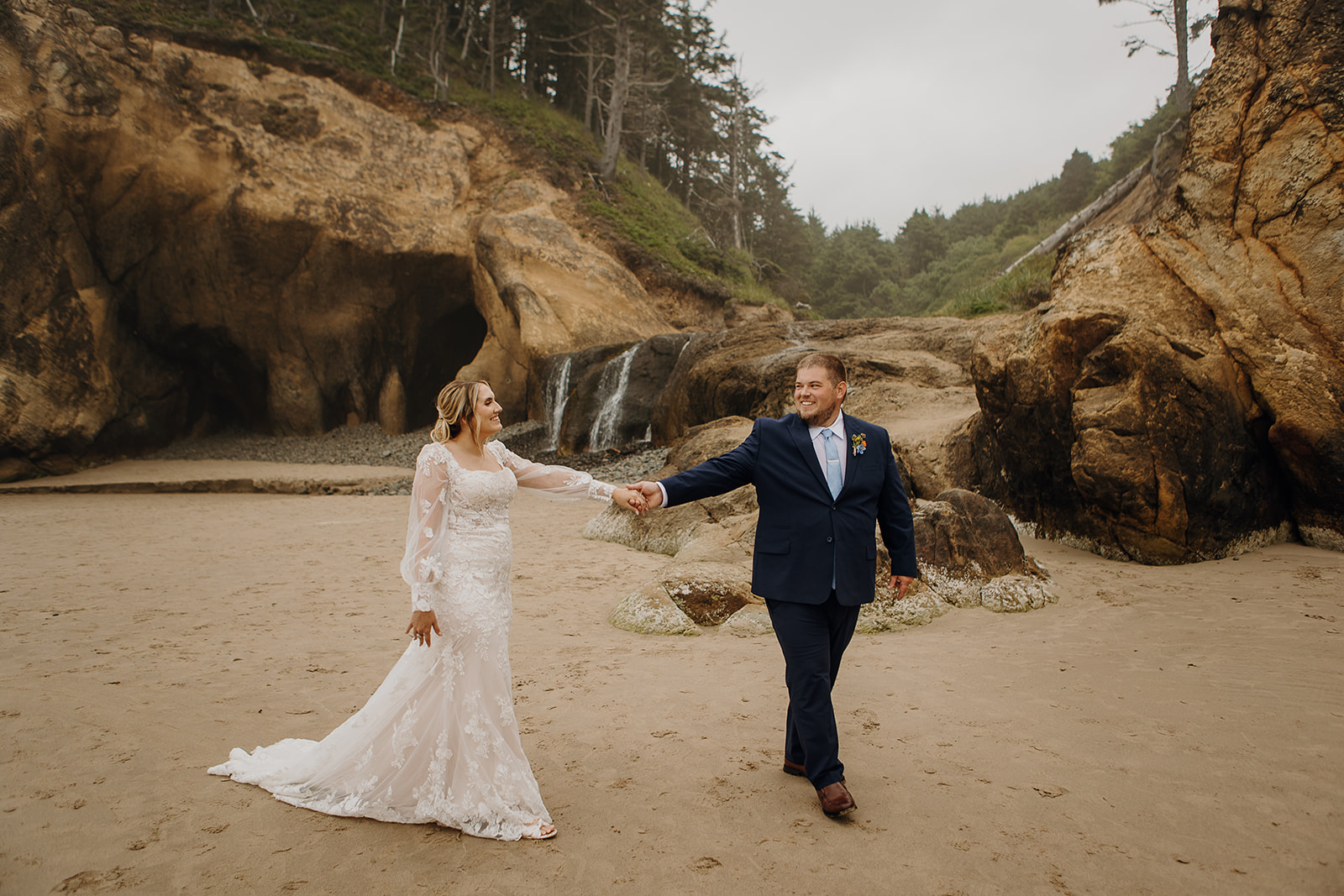 a man and woman in wedding gear walk hand in hand in front of the waterfall at Hug Point Falls on the Oregon Coast.