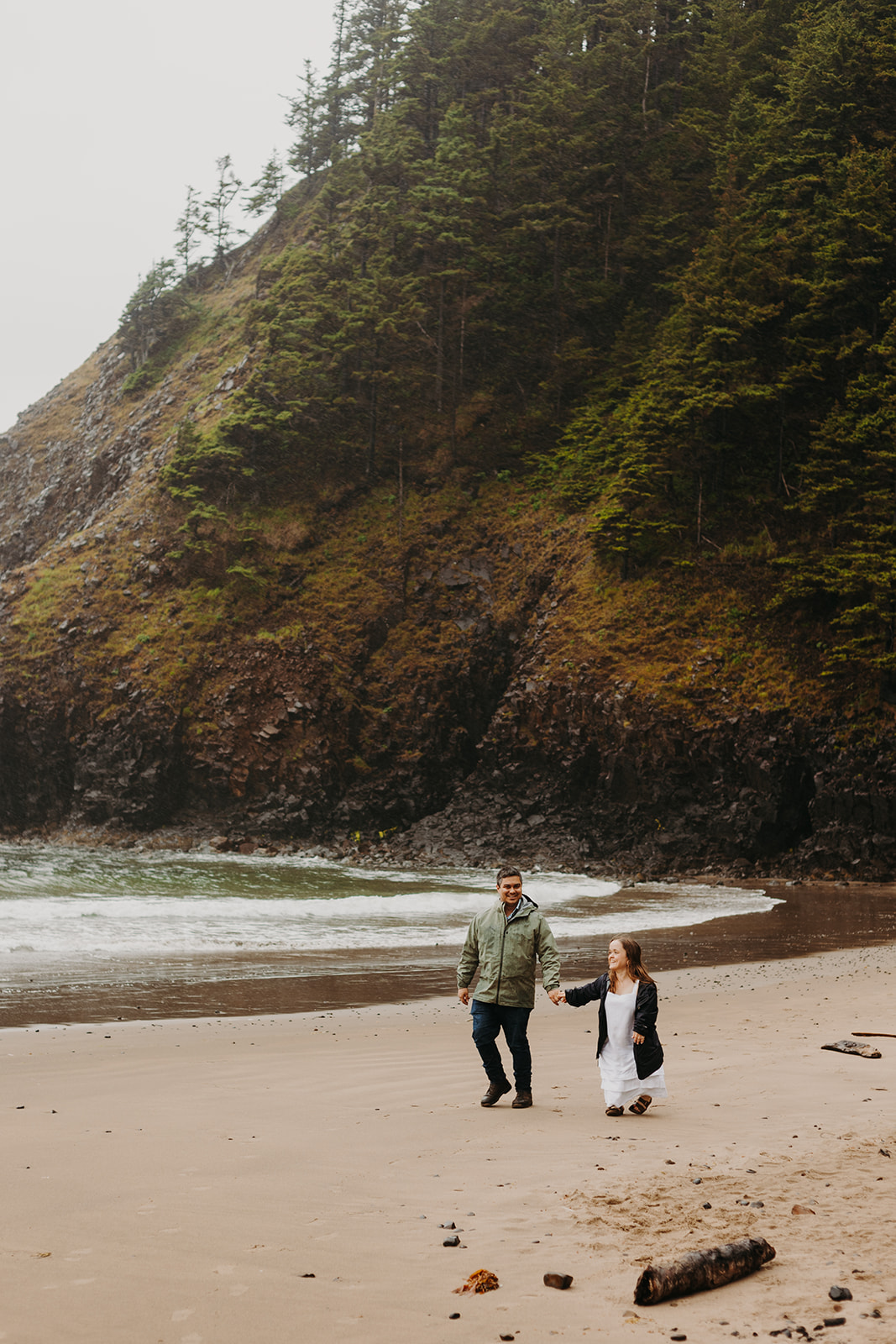 man and woman walk hand in hand in the rain on Indian Beach with a forested cliff and ocean in the background
