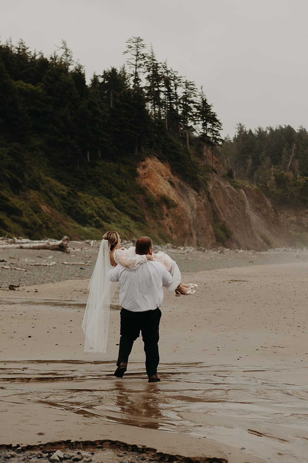 a groom walks through a puddle on the beach carrying his bride in his arms