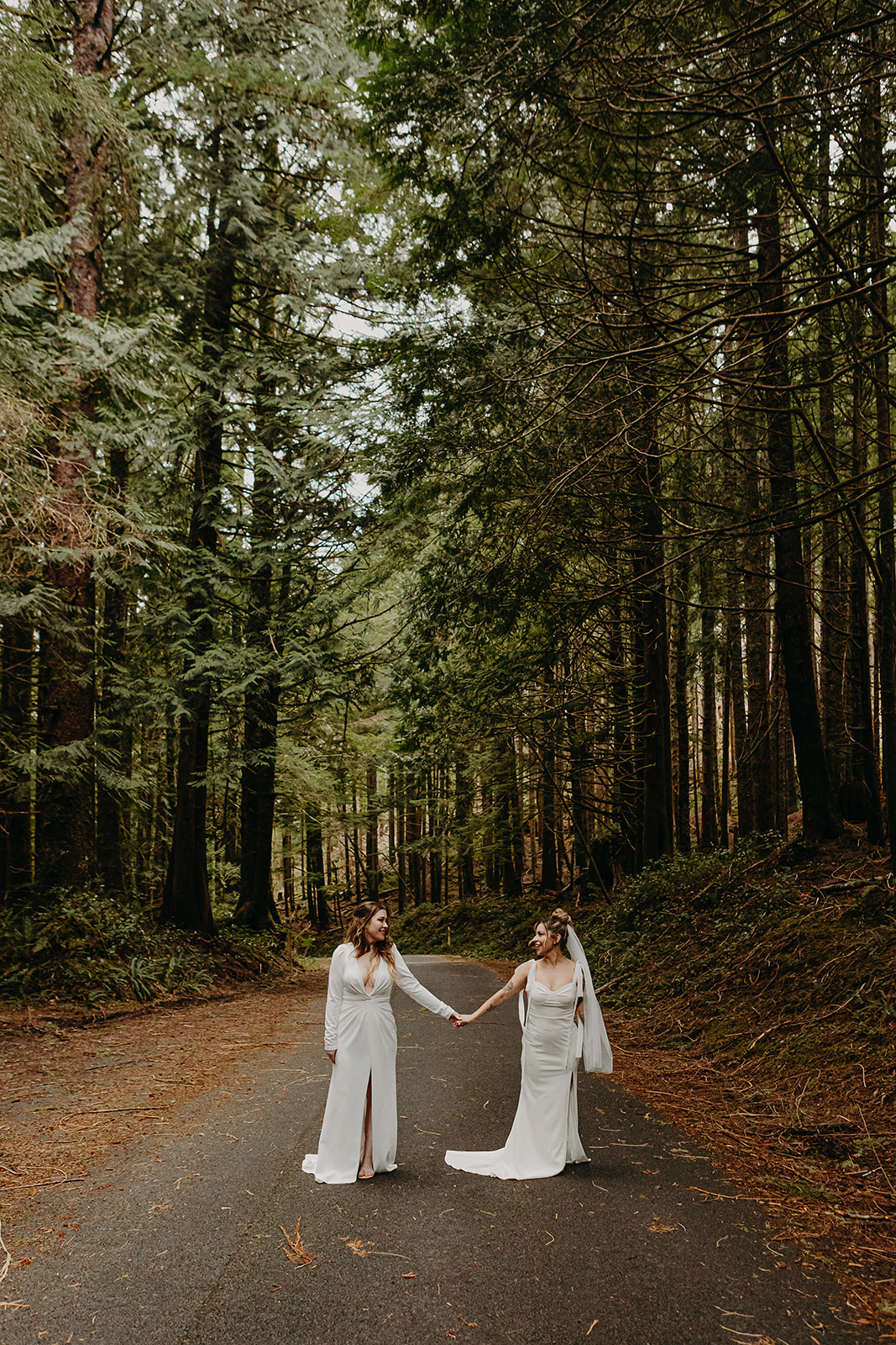 two brides stand hand in hand in a forest in the middle of a trail road