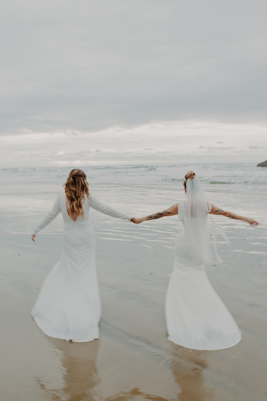two brides run hand in hand on a beach into the waves at Manzanita Beach in Oregon