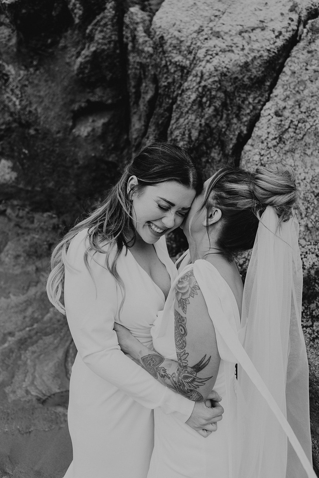 two brides stand on a beach as one whispers excitedly into the other's ear