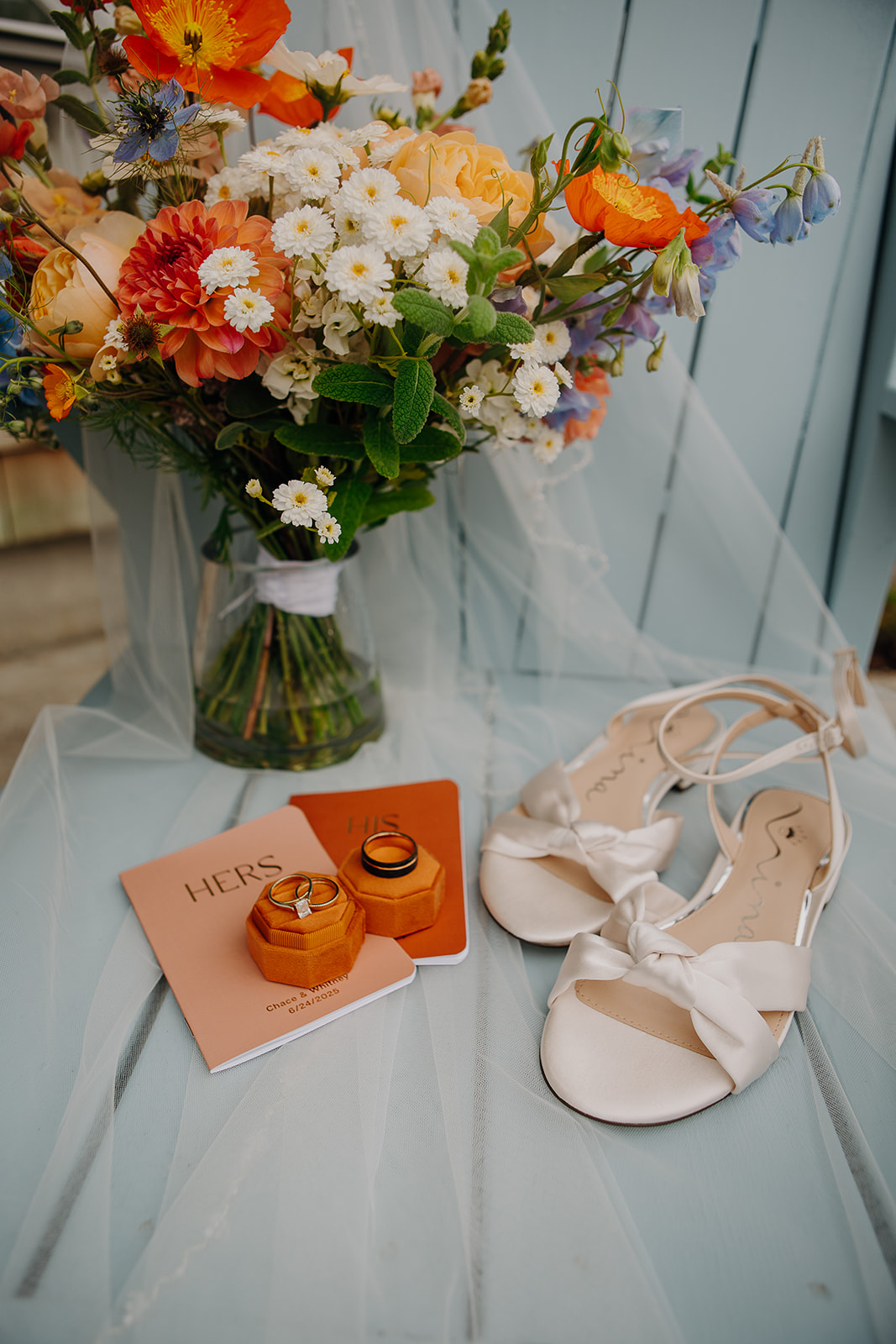 a blue and orange wedding bouquet sits on an orange chair with wedding vow books, rings, and wedding shoes