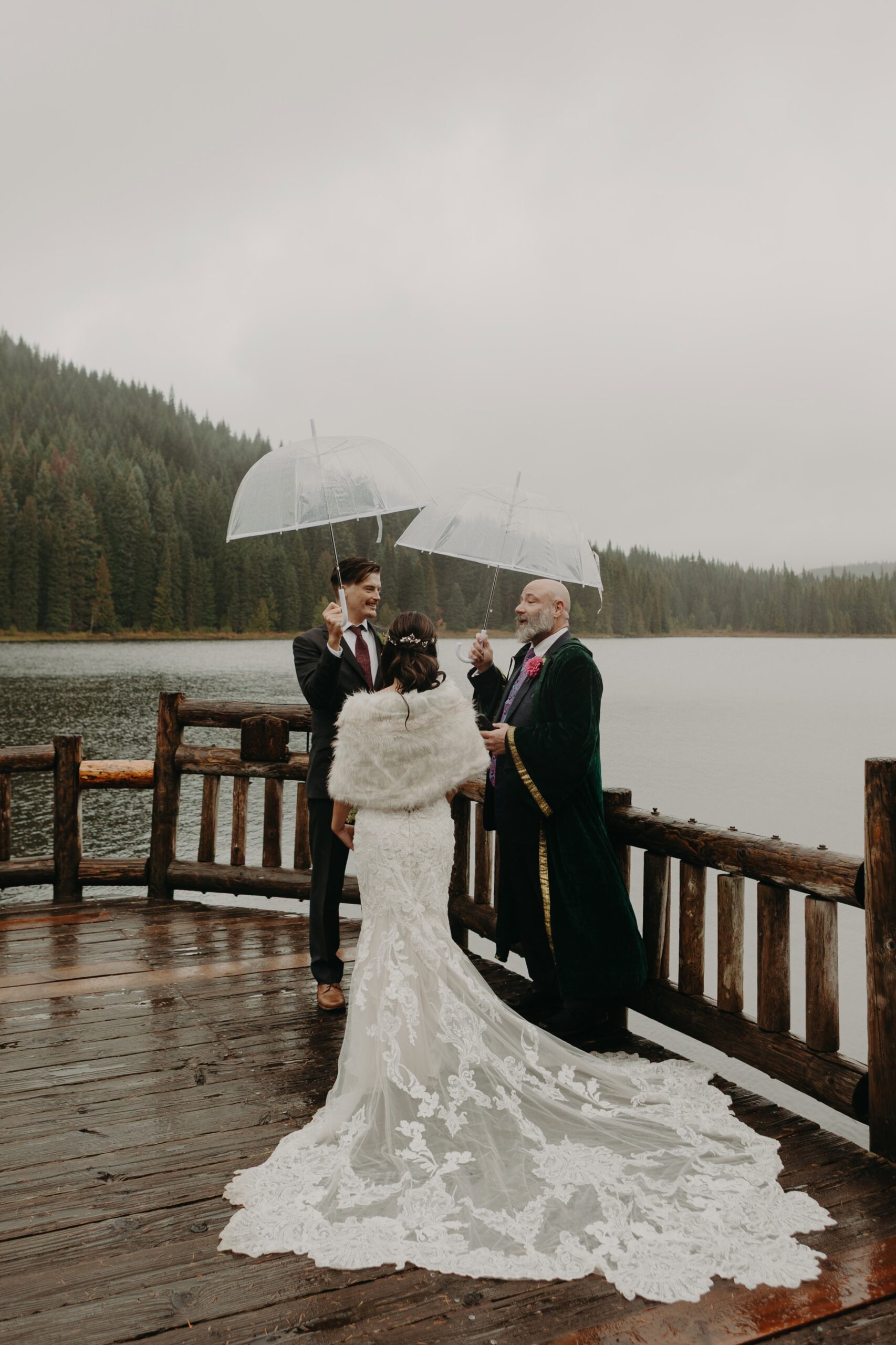 an officiant stands before a bride and groom holding umbrellas on the dock at Trillium Lake