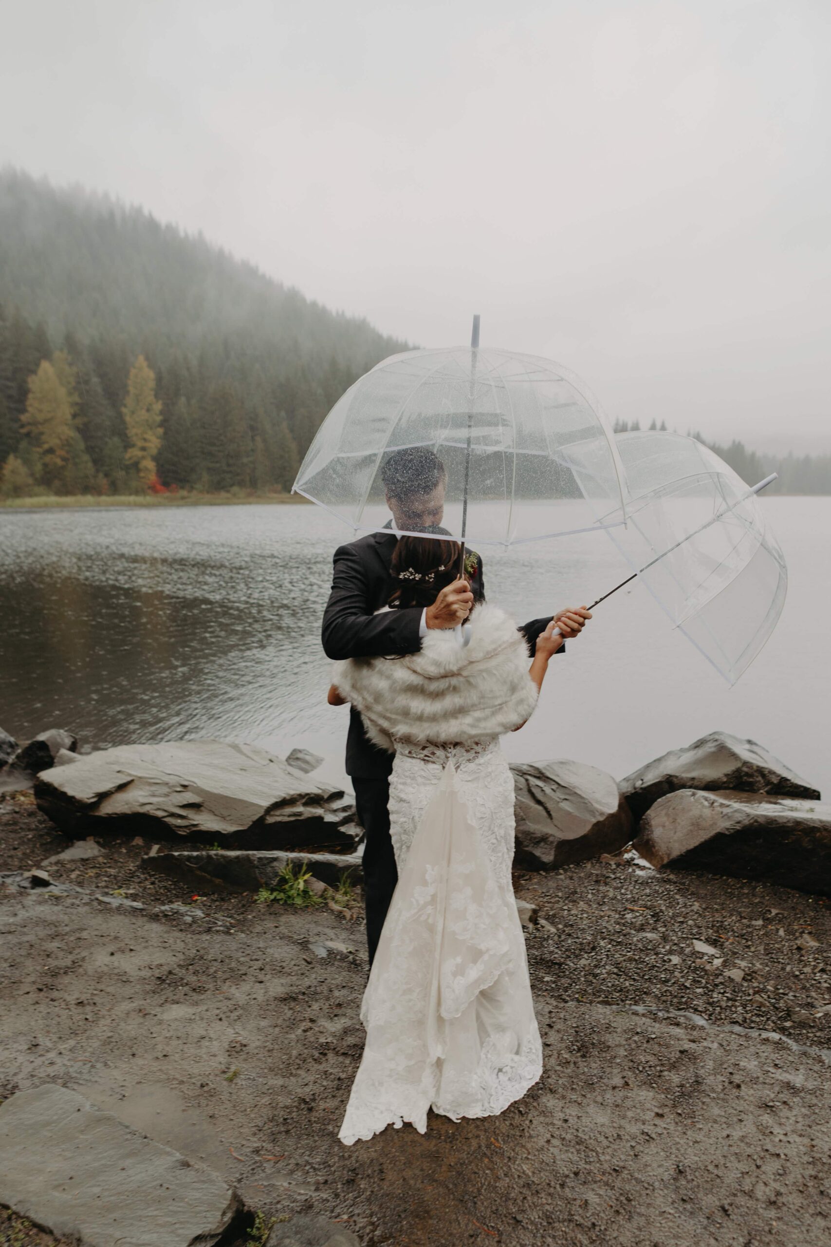 bride and groom embrace holding clear umbrellas at the shore of Trillium Lake 