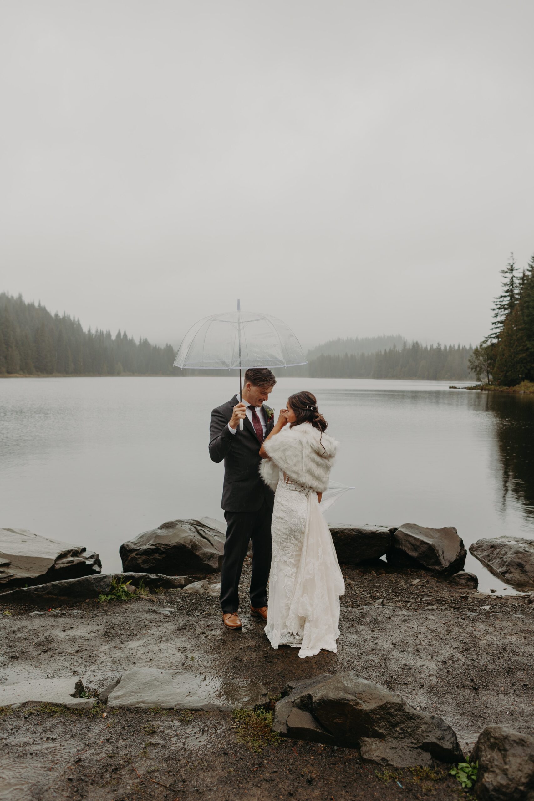 bride and groom shed a happy tear for their first look at mount hood's trillium lake while they both hold umbrellas