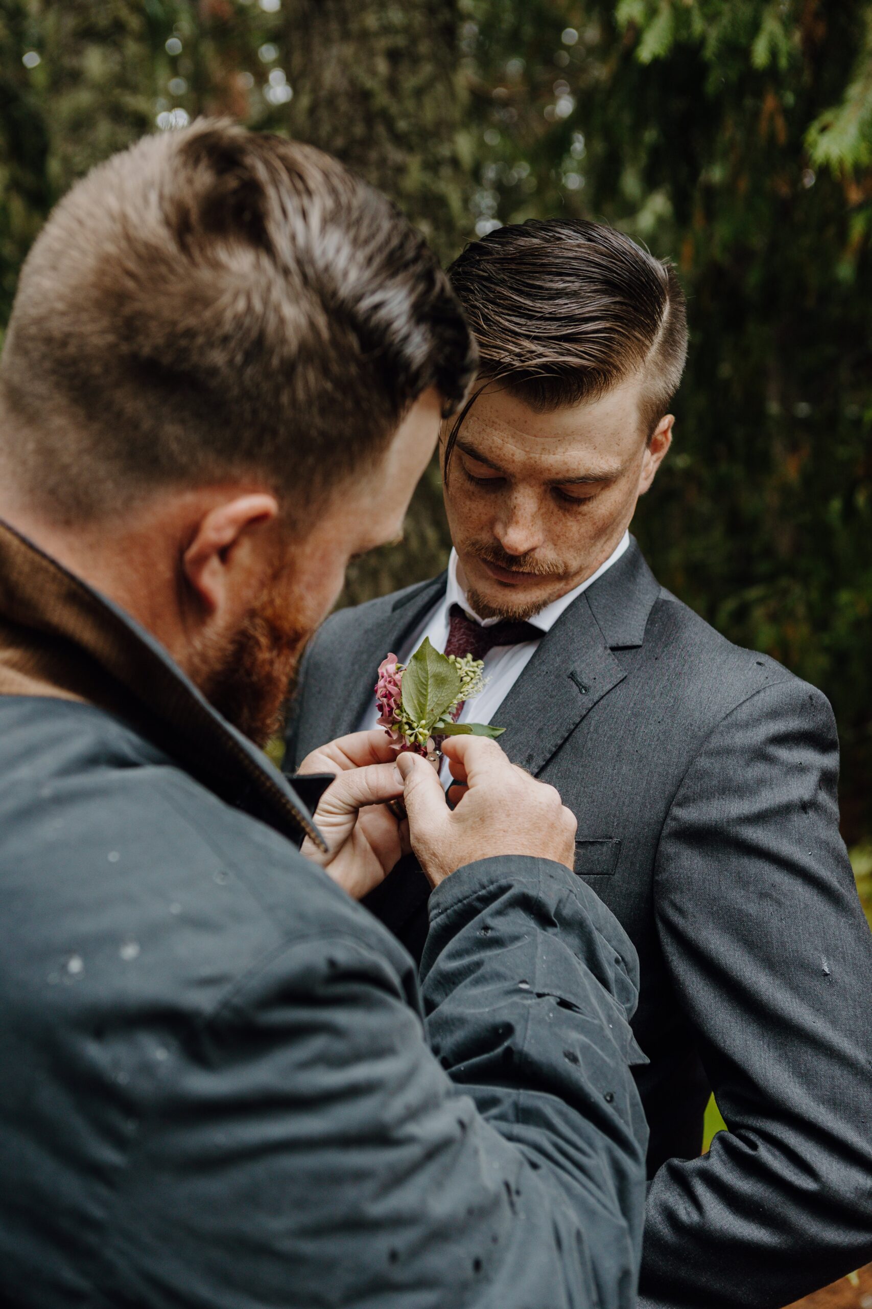 groomsman pins boutonniere on groom outside surrounded by trees as rain sprinkles down