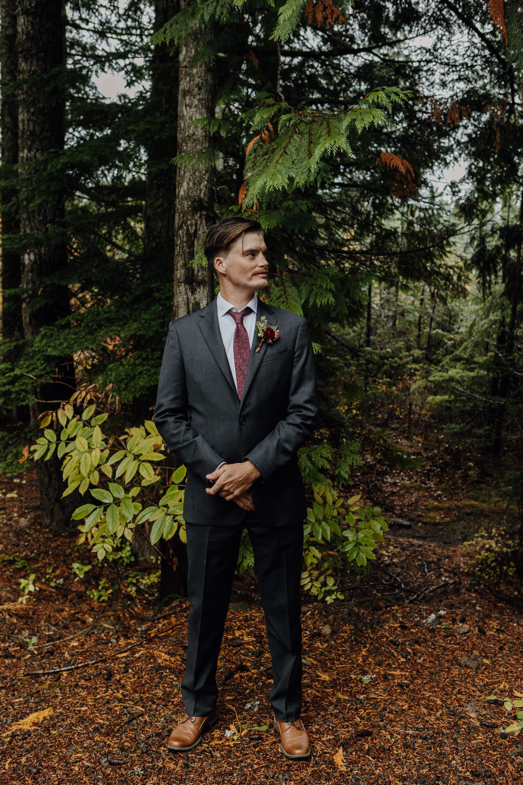 a groom stands in his suit in a forest of lush greenery holding his hands