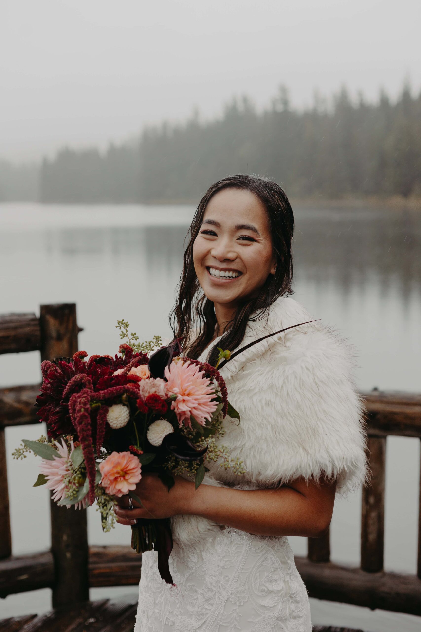 bride stands on a pier with wet hair but still smiling happily holding a wedding bouquet of pink and deep red flowers