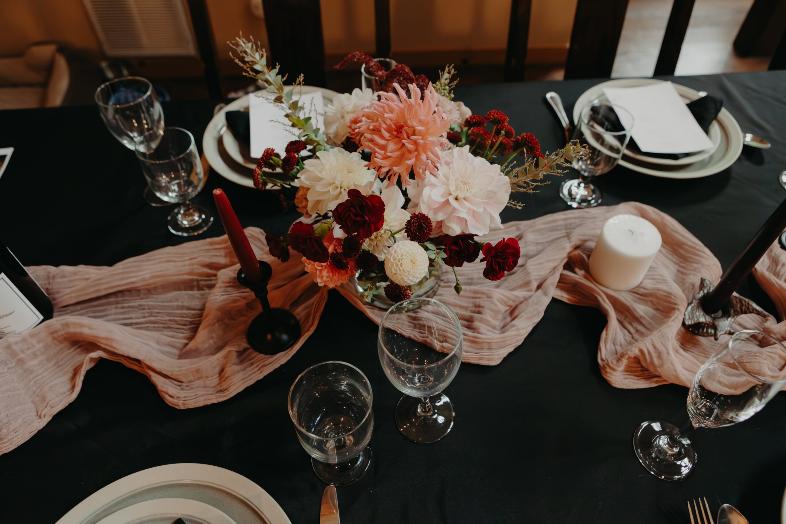 pink table runner on black tablecloth with floral centerpiece of pink and maroon flowers, surrounded by cutlery and plates for a wedding dinner