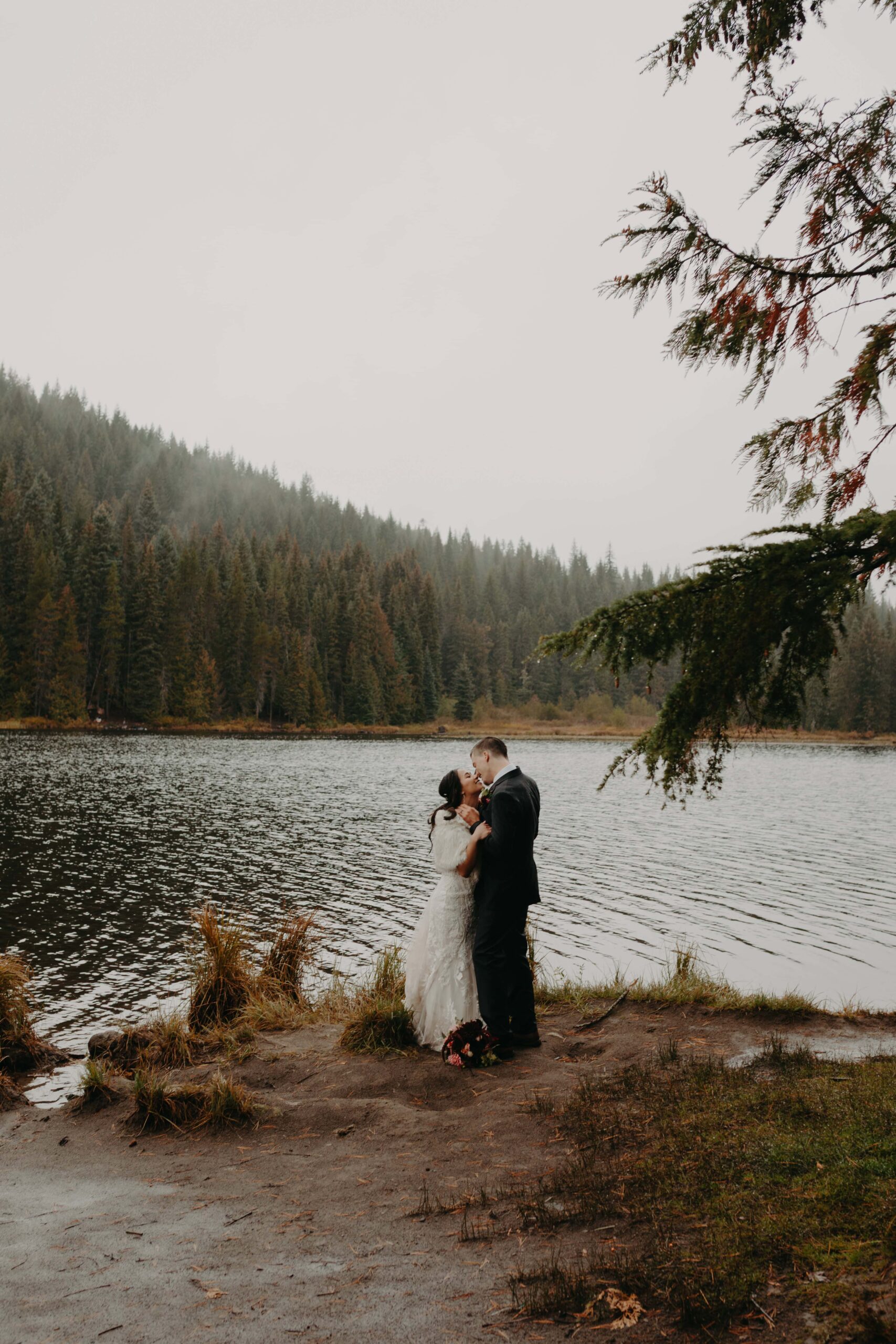 bride and groom stand on the shores of trillium lake embracing with mist and clouds in the distance atop the trees