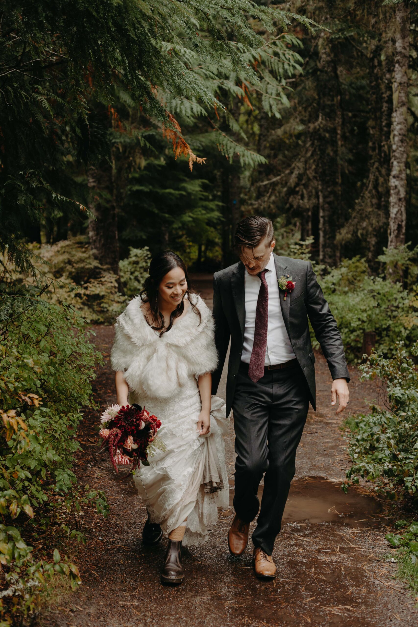 groom walks side by side with his bride holding her train as they walk through a forested trail