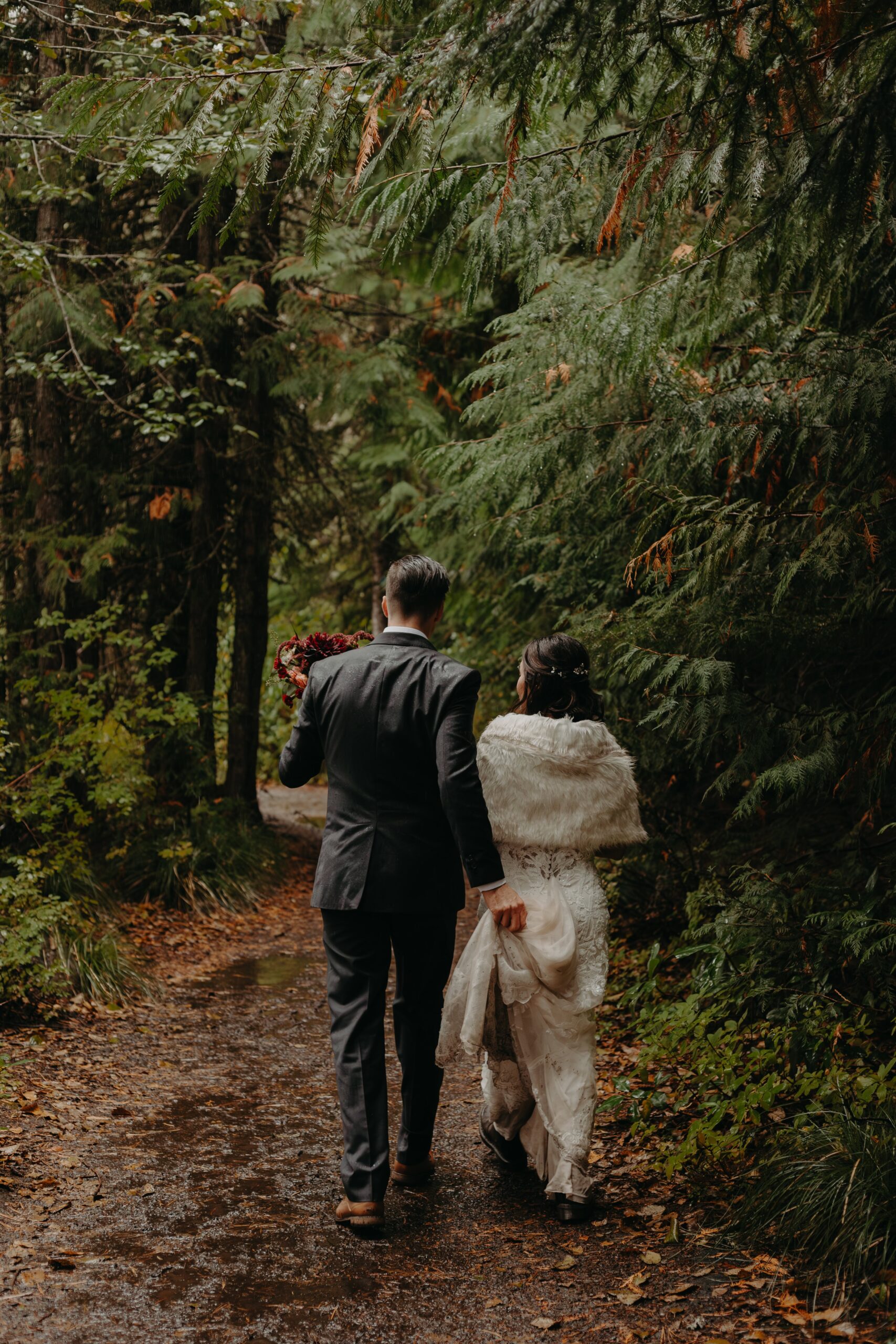 groom walks side by side with his bride holding her train as they walk through a forested trail