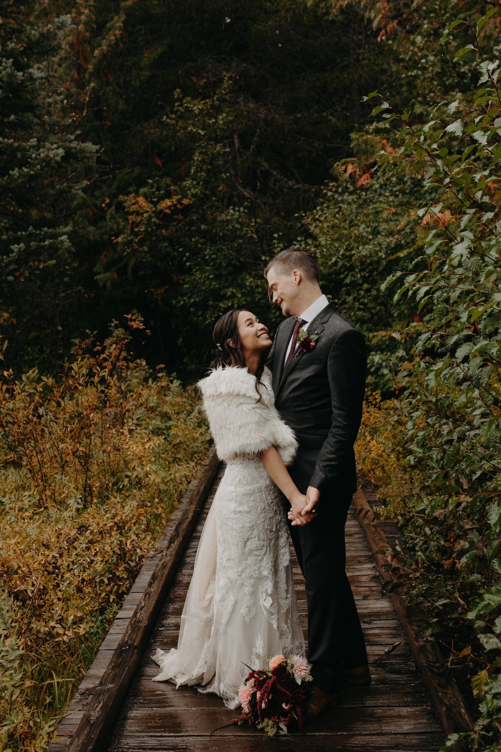 bride and groom stand hand in hand on a wooden bridge with autumnal trees in the background